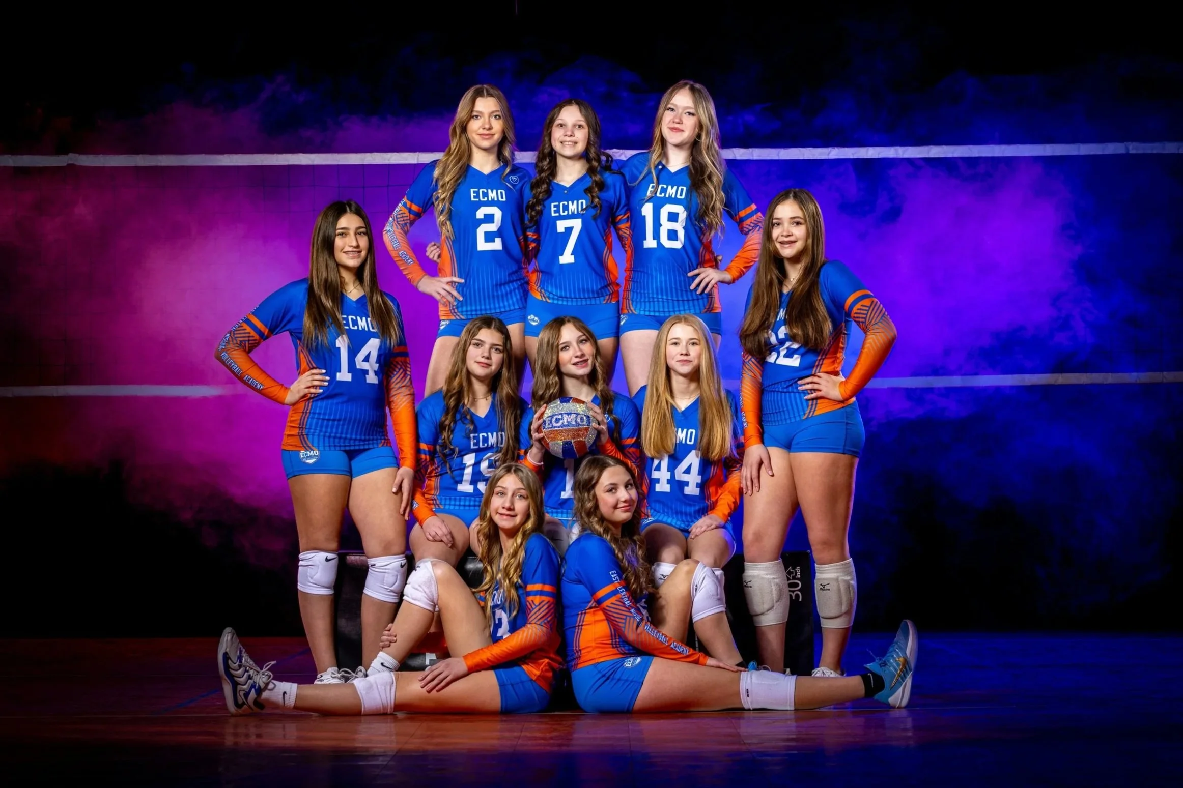 A girls' volleyball team wearing blue and orange jerseys, posing on a court with a purple and blue background. They are arranged in a group with some standing, some kneeling, and one sitting on the floor, holding a volleyball.