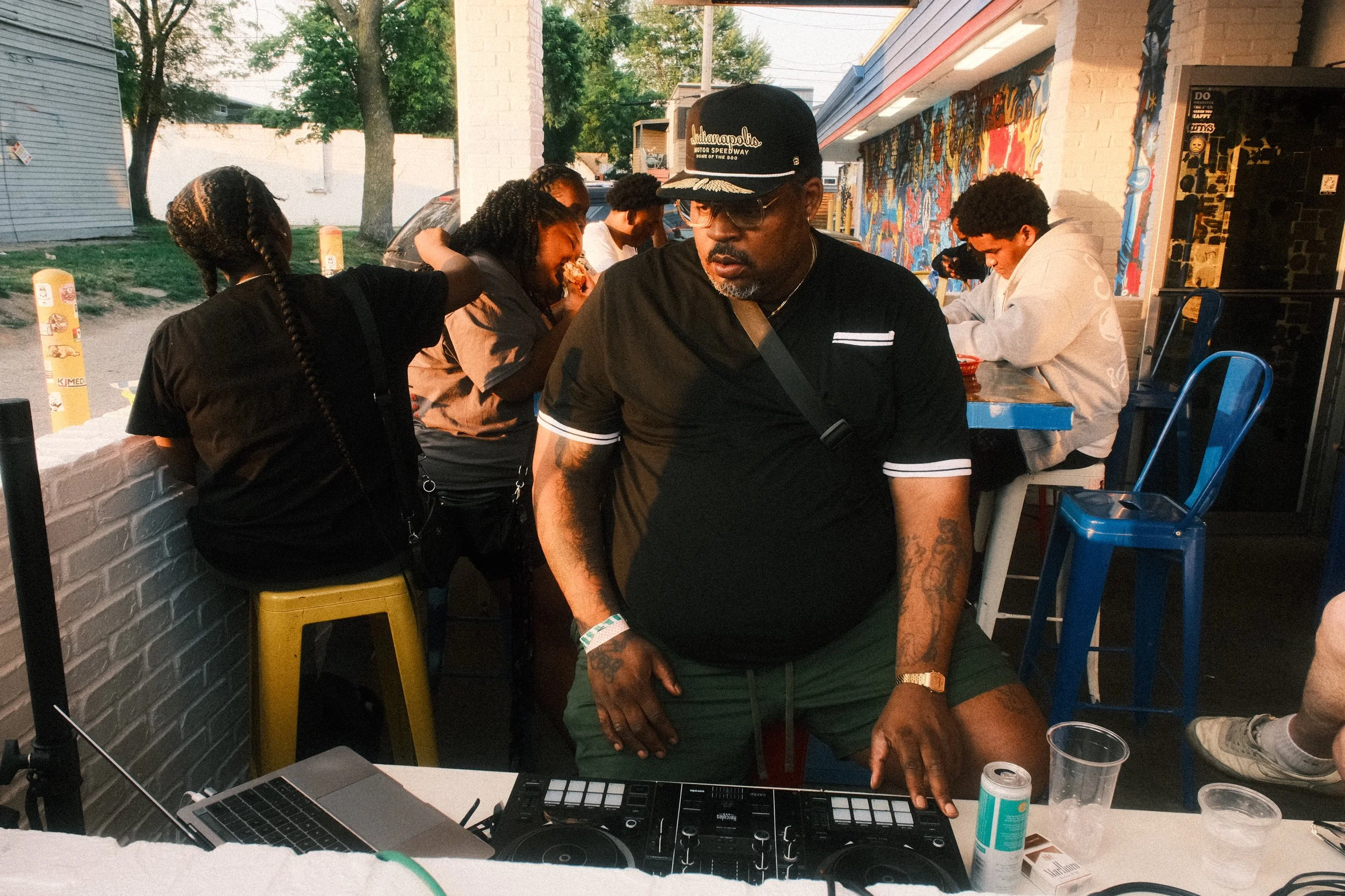 A man wearing glasses, a black t-shirt, a black cap that says 'Indianapolis Motor Speedway,' and a gold watch is DJing with a music controller at an outdoor gathering. Several people are around him, some sitting and others standing, with one woman leaning in close to talk to another person. People are casually dressed, and there is a colorful mural on the wall behind them. The scene appears to be at dusk with trees and buildings in the background.