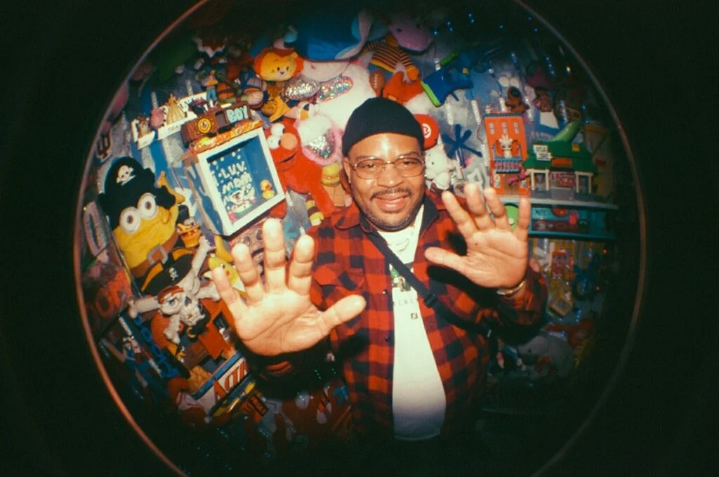 A man in a red and black plaid shirt and glasses stands with his hands raised in front of a colorful background filled with plush toys and toys, smiling at the camera.