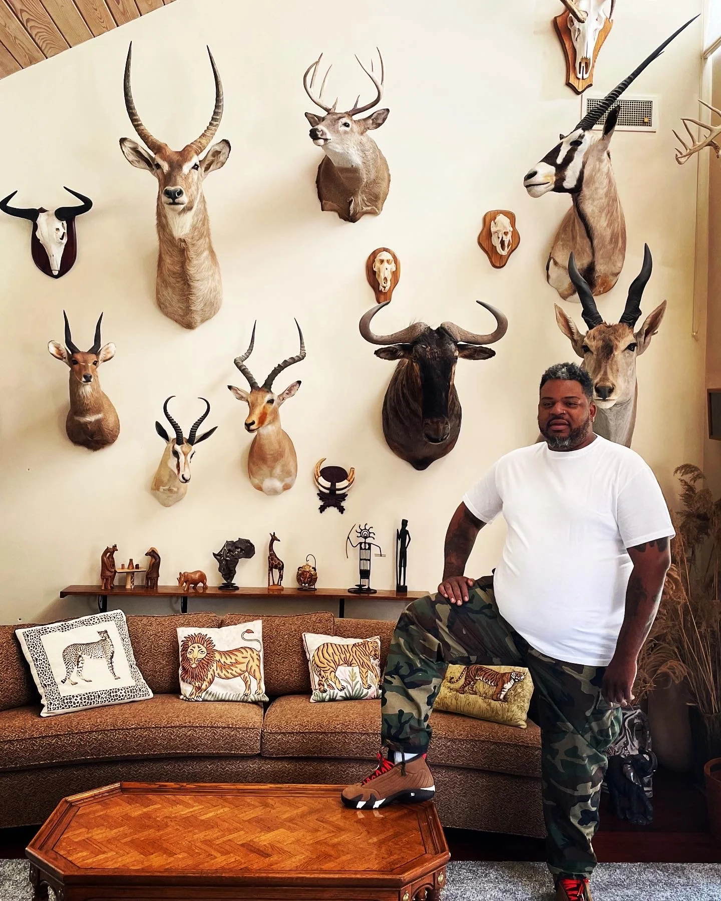 A man with short curly hair, a beard, wearing a white T-shirt, camouflage pants, and tan sneakers, standing in a room with a wall full of mounted animal heads, including deer, antelope, and a unicorn. There is a brown sofa with lion-themed pillows and a wooden coffee table in front.