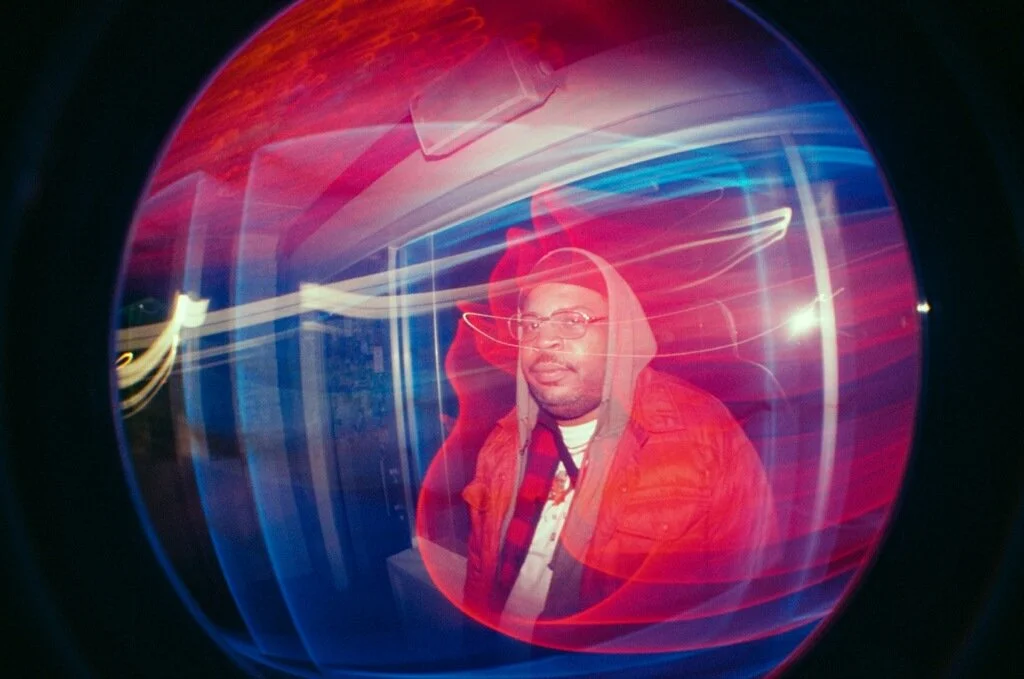 A person in glasses wearing a hoodie and jacket looking through a colorful, distorted glass or lens with red, blue, and purple light streaks.