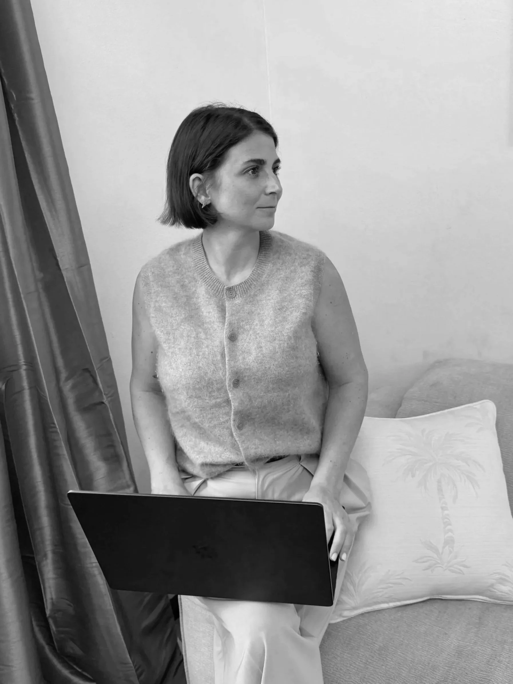 A woman with short dark hair, wearing a sleeveless cardigan and earrings, is taking a mirror selfie in a room with plain walls and a bed with a patterned pillow. She is holding a smartphone in her right hand.