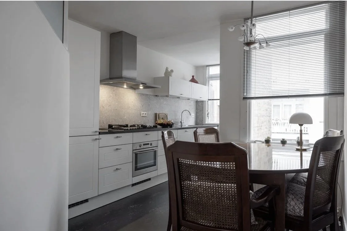 Modern kitchen with white cabinets, stainless steel stove and oven, gray backsplash, and a dining table with four wooden chairs. Large windows with blinds let in natural light.