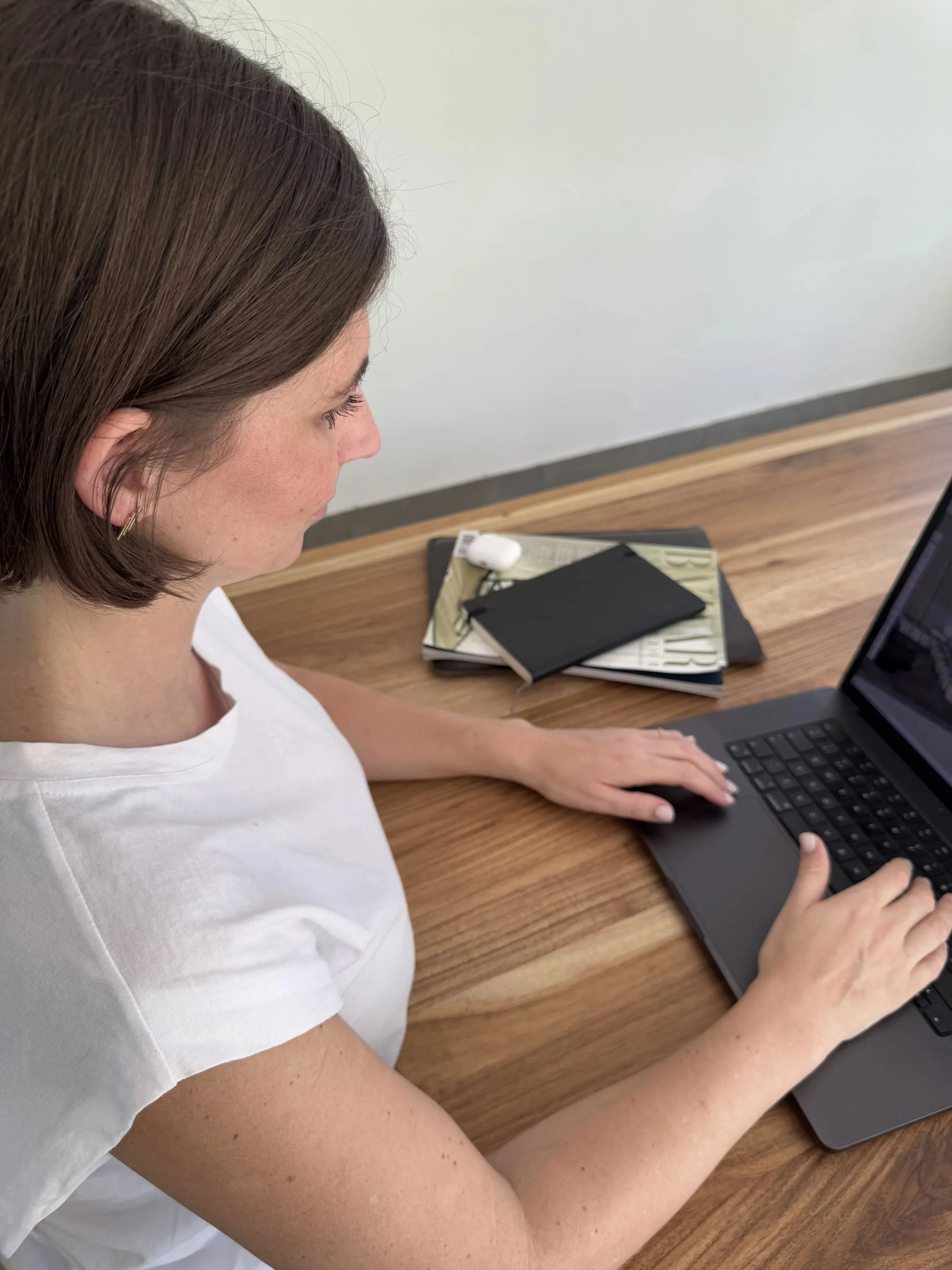 A woman with brown hair and light skin wearing a white T-shirt sitting at a wooden desk working on a black laptop with a black notebook, a white earbud case, and printing materials nearby.