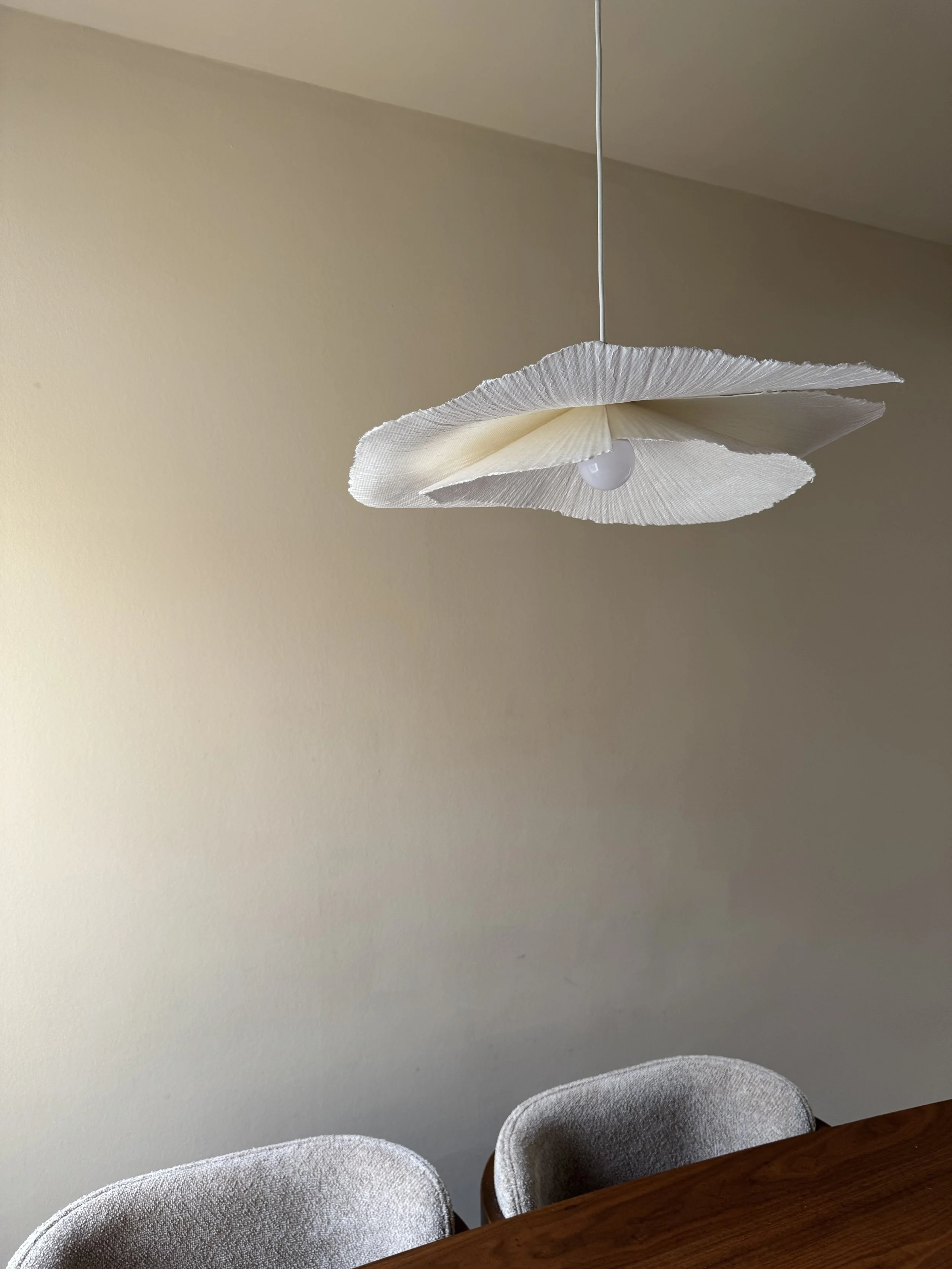 A hanging white paper lampshade with a light bulb inside, above a wooden dining table surrounded by gray upholstered chairs in a room with beige walls.