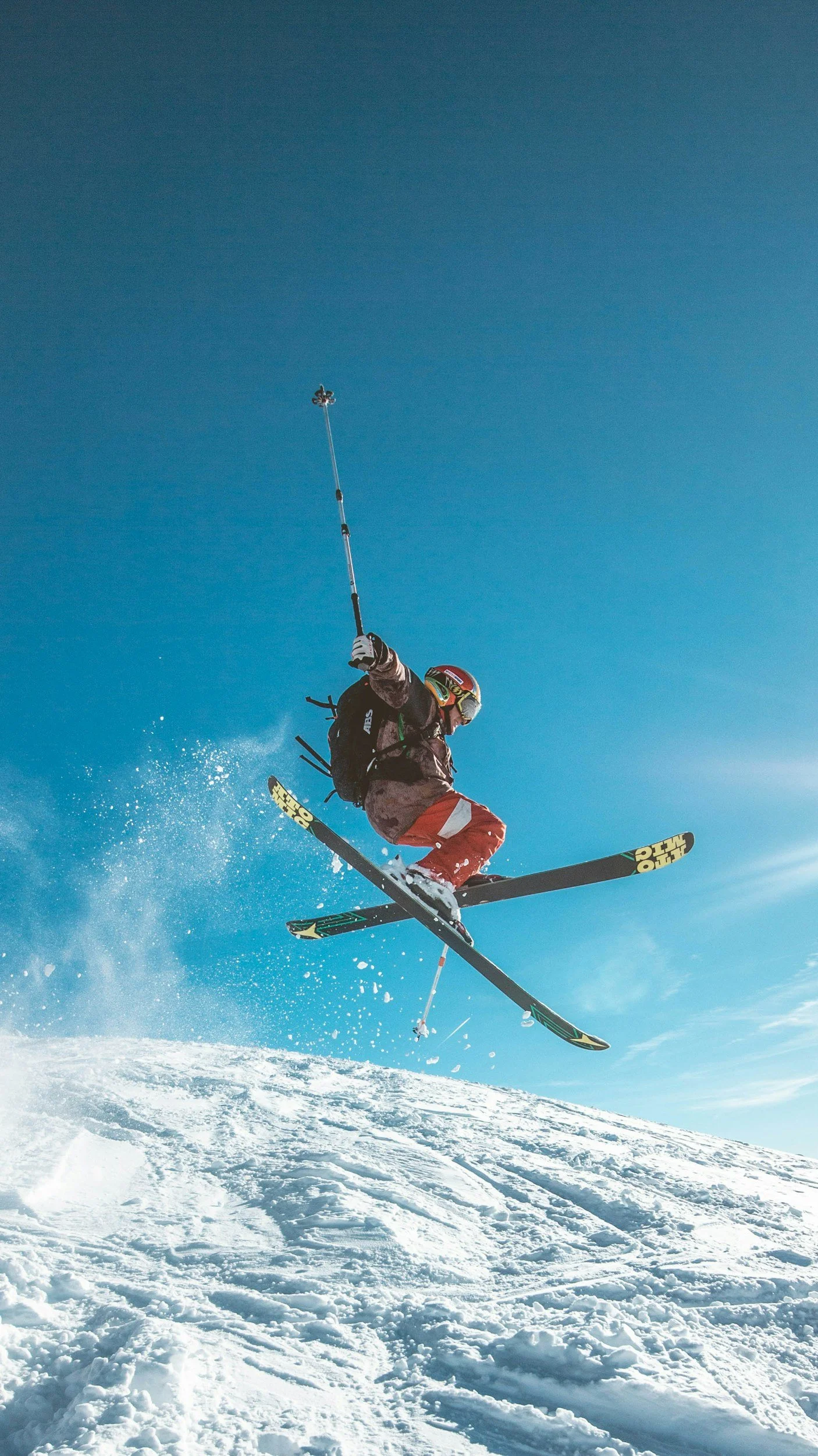 A skier in mid-air performing a jump on a snowy mountain under a clear blue sky, wearing a helmet, goggles, and winter gear.