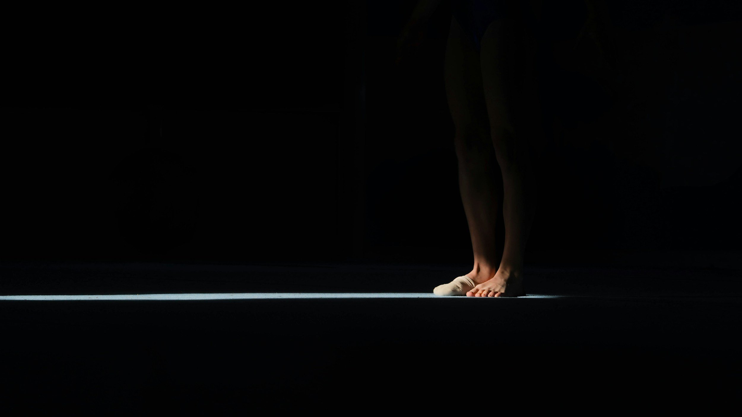 A dancer's feet in ballet slippers, standing on a stage with a dark background and white line, with the upper body out of frame.