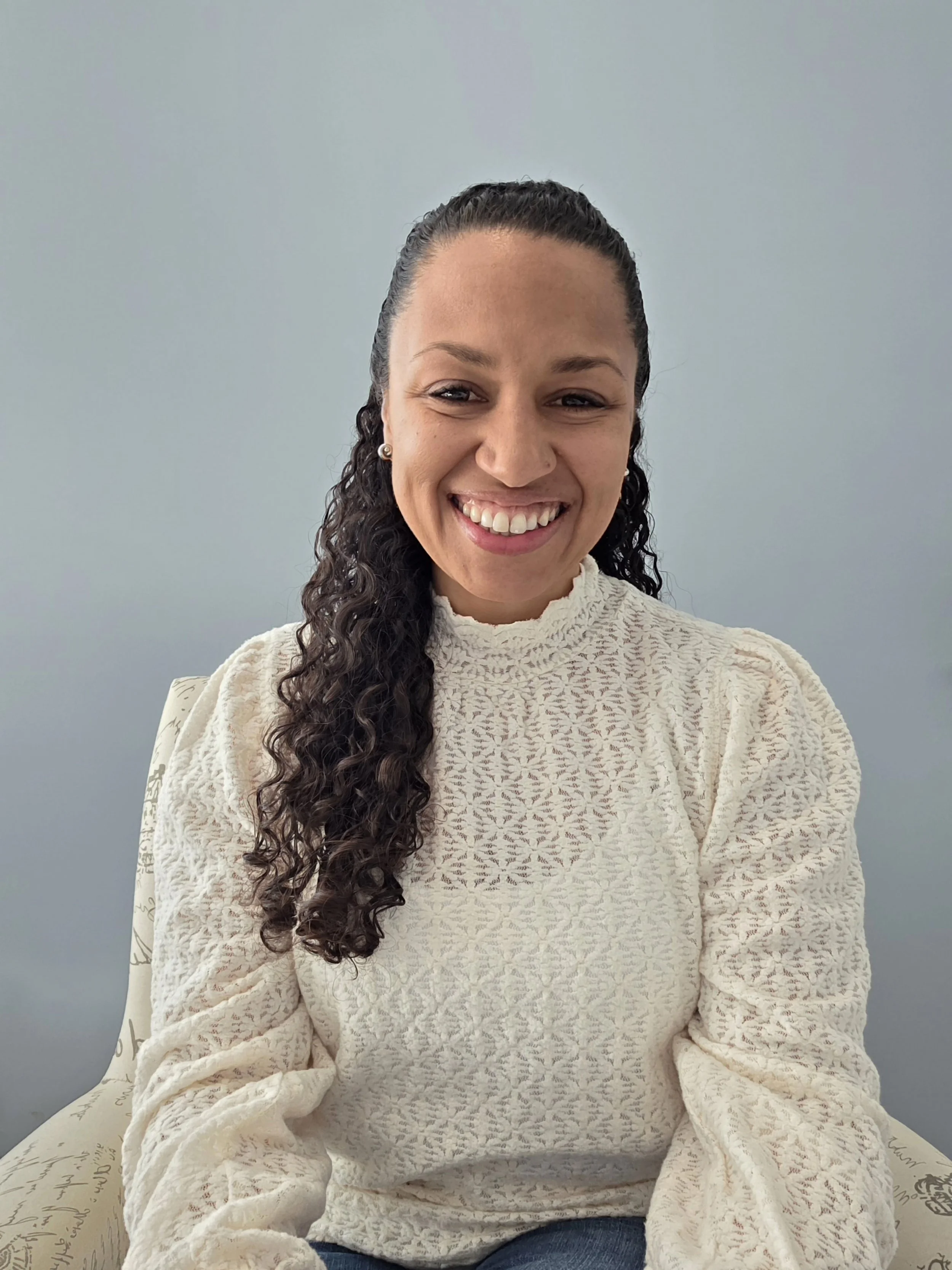 A woman with curly dark hair, wearing a white lace top, sitting on a chair with a patterned cushion against a light gray wall. She is smiling.
