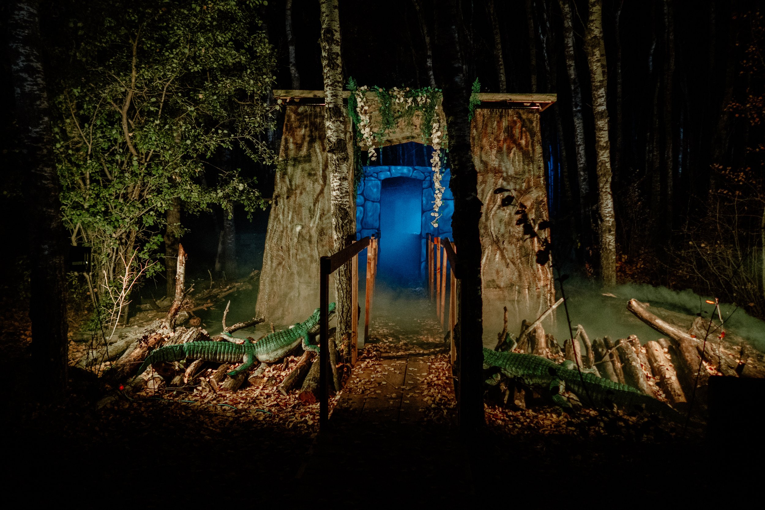 A nighttime scene of a forest with trees and a stone archway decorated with artificial leaves and flowers, illuminated with blue lighting.