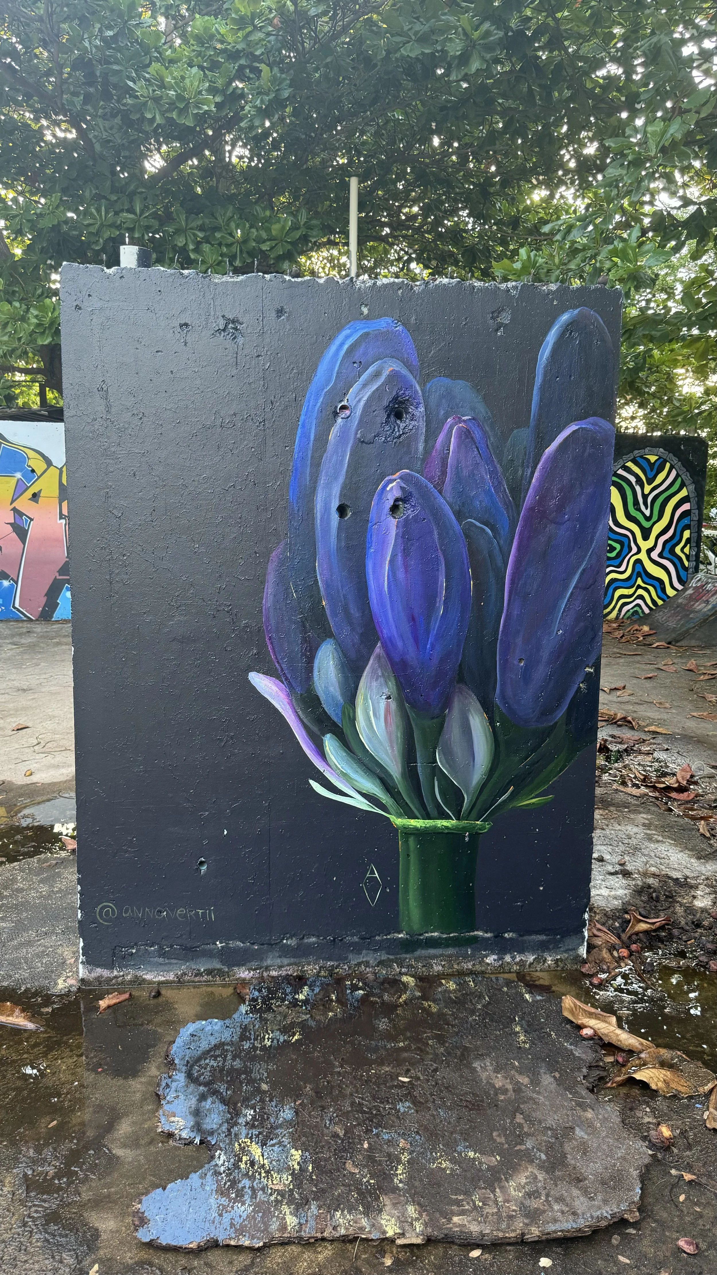 AnnaV mural in Puerto Rico — moody purple flower with thick painterly petals emerging from a green stem, on a black wall in a street-art park