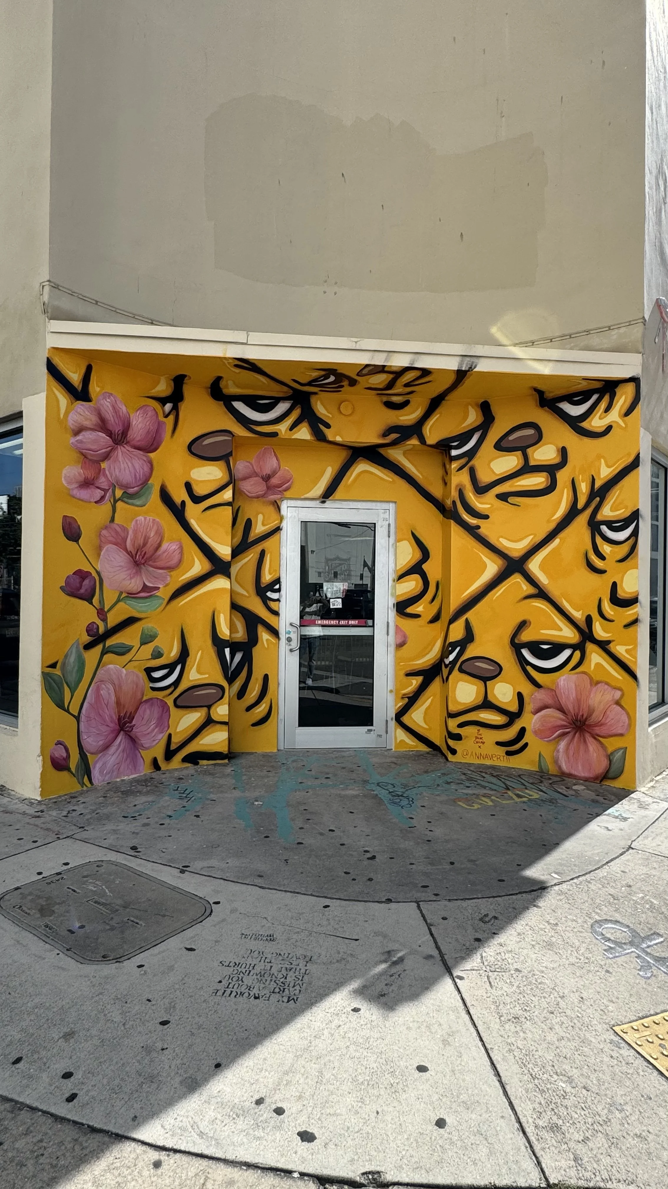 Mural by AnnaV and JC Rivera in Miami Wynwood — JC Rivera's yellow bear characters in a black diamond grid, with AnnaV's pink and coral florals climbing through, wrapped around a building entrance