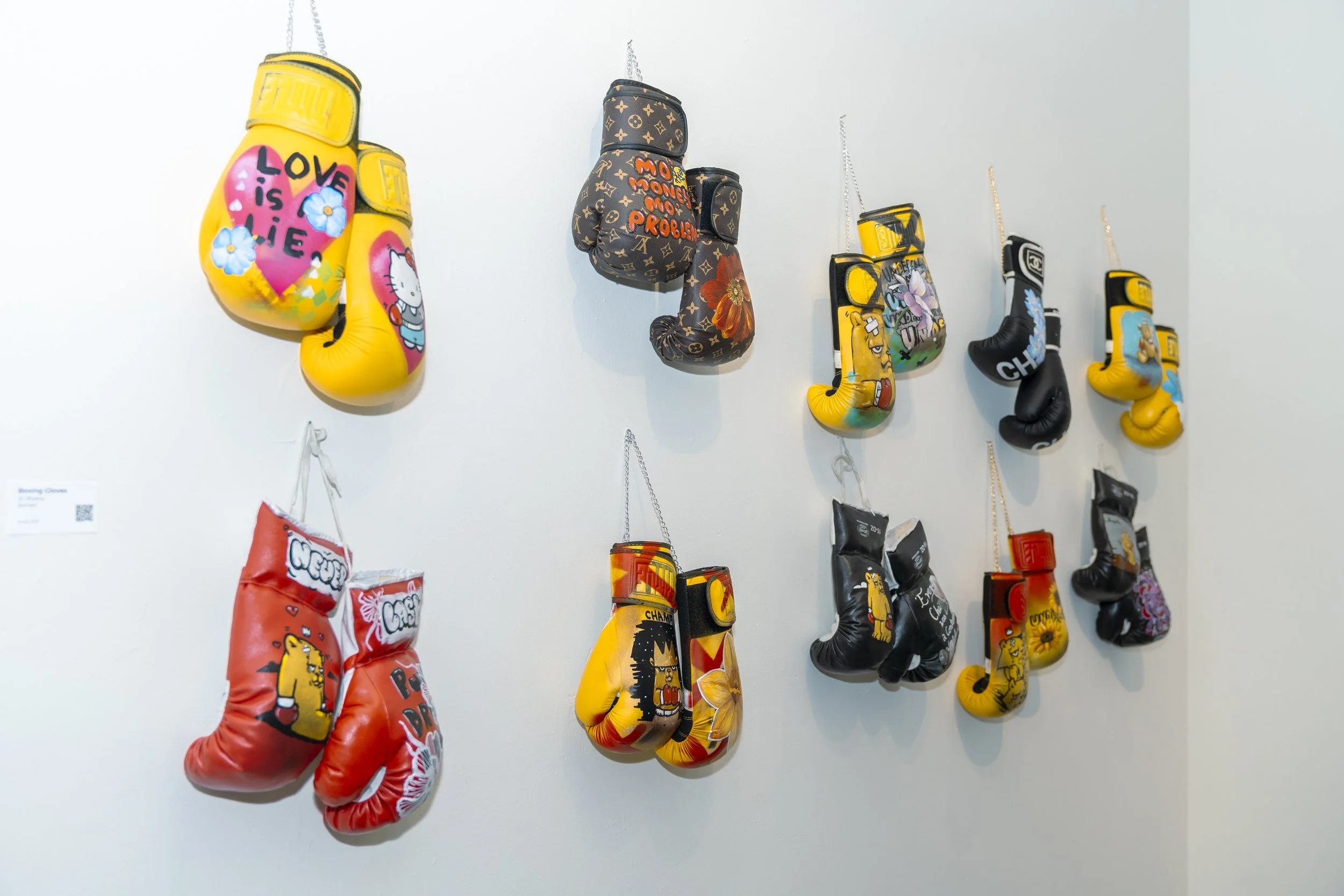 AnnaV and JC Rivera collaboration — wall display of hand-painted boxing gloves hung by their laces, each one customized with florals and characters, at Common Ground exhibition