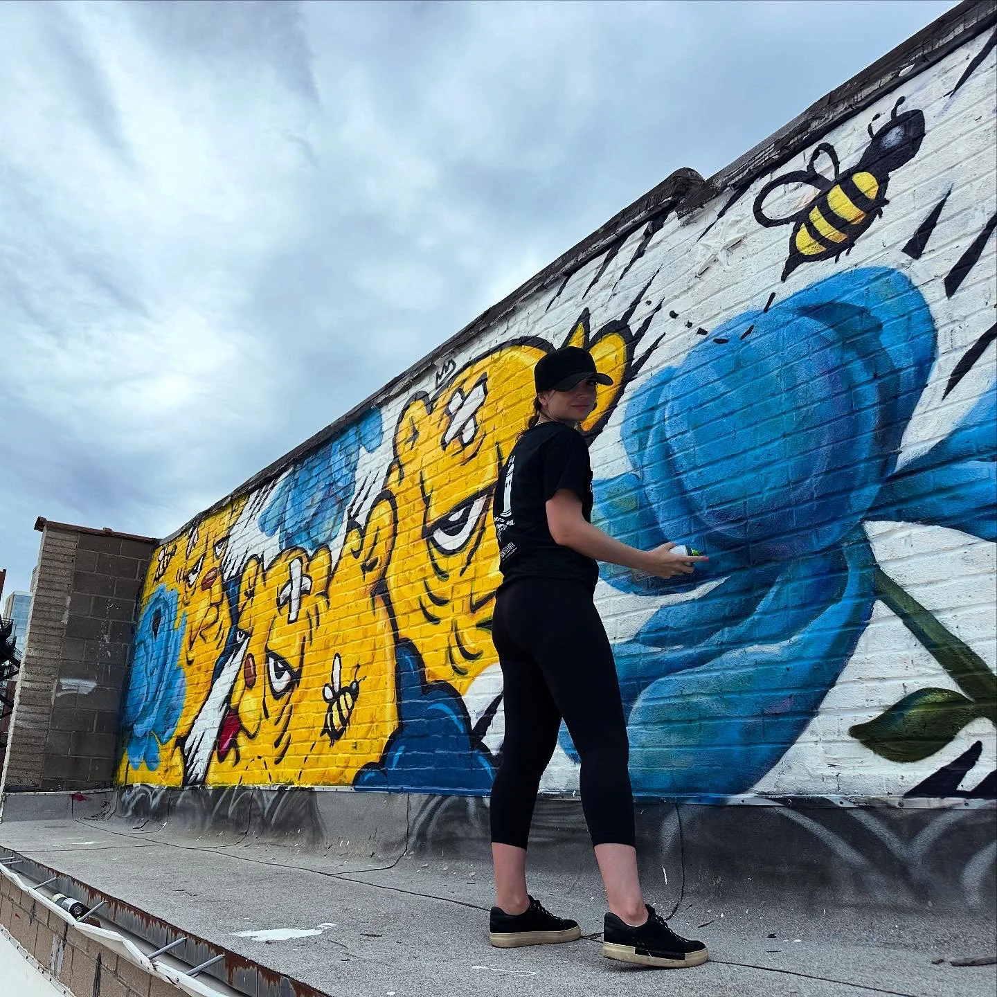 AnnaV at work on a Kansas City mural, spray-painting a blue peony into a wall of yellow bear faces and bees — collaboration with JC River