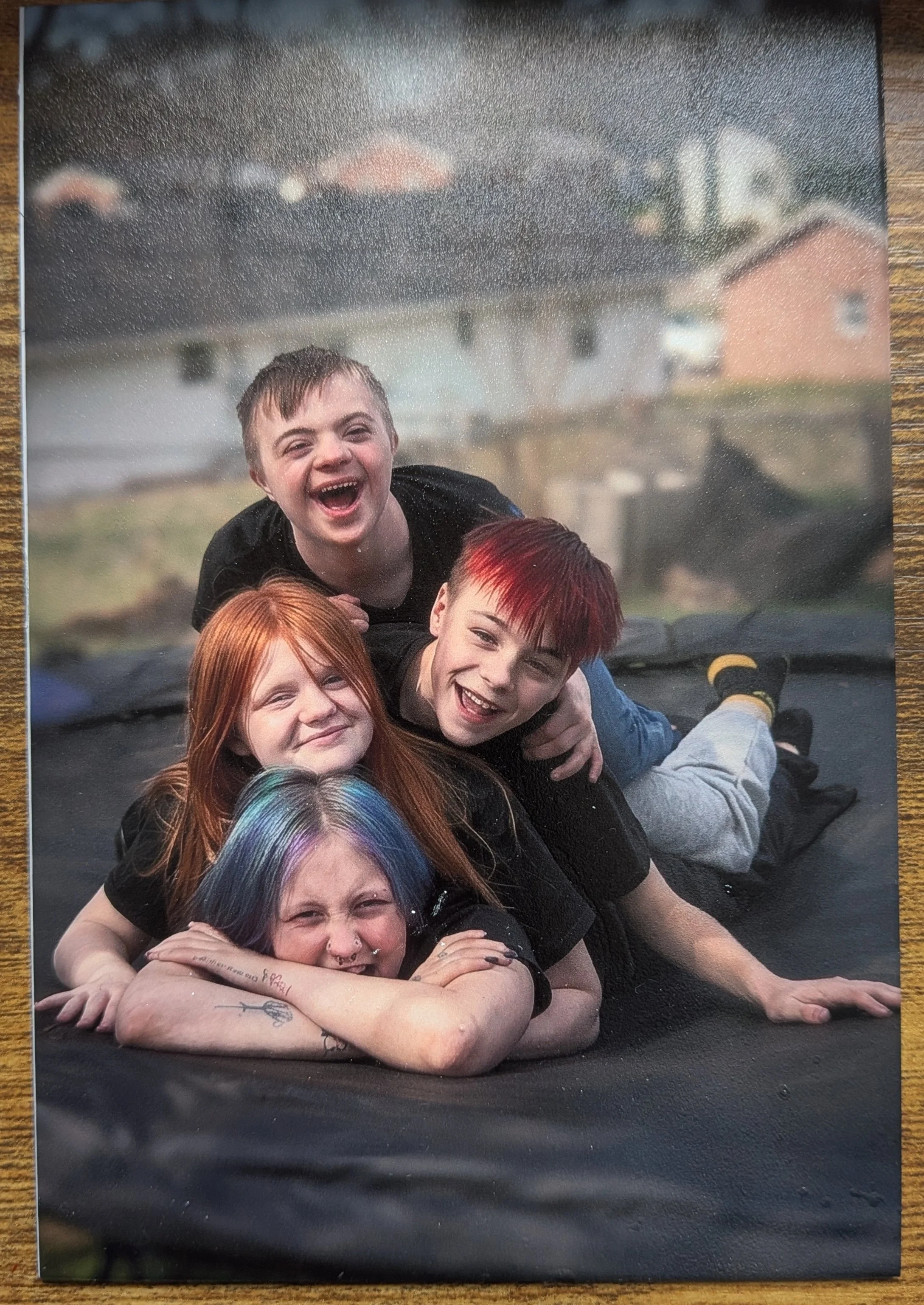 A group of young people, including boys and girls, are stacked on top of each other, smiling and laughing while laying on a large black trampoline outdoors. UV Printed