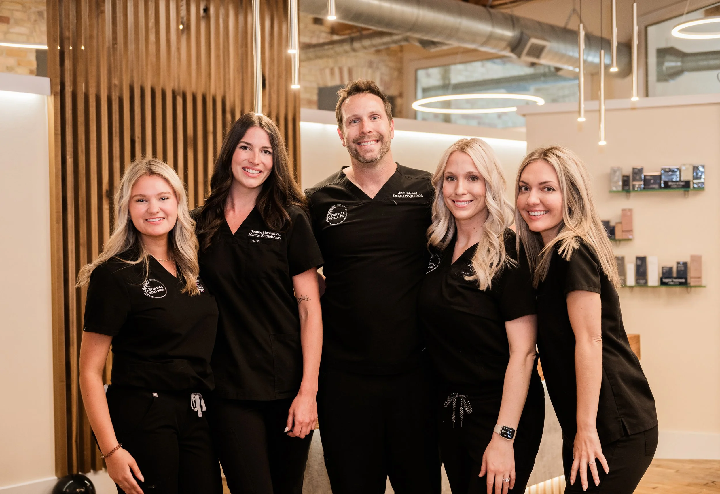 Group of five smiling healthcare professionals, four women and one man, standing indoors in a modern clinic or wellness center, wearing black scrubs with embroidered logos.
