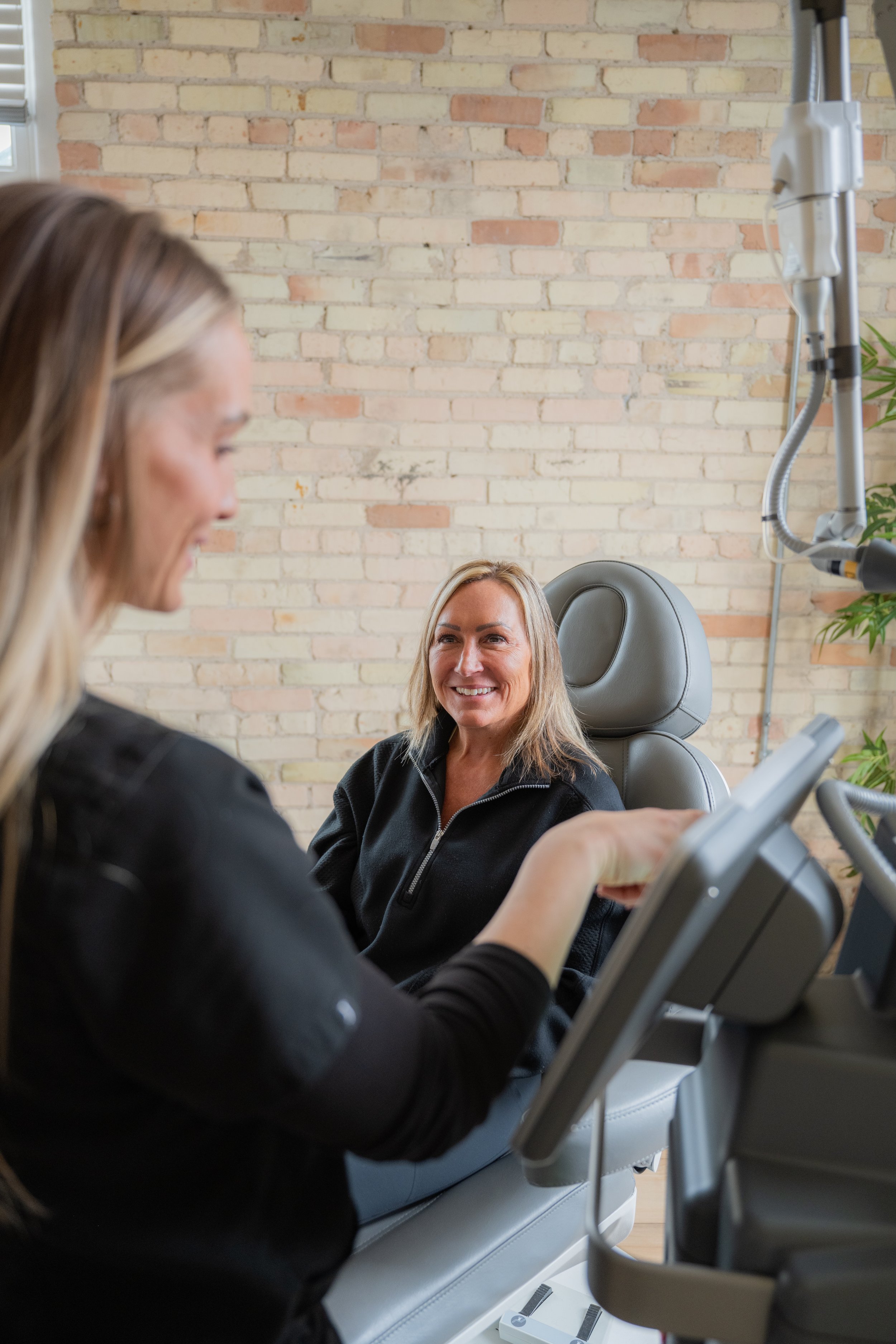 A woman is sitting in a mespa chair, smiling, while a medspa professional is examining her face and holding an instrument.