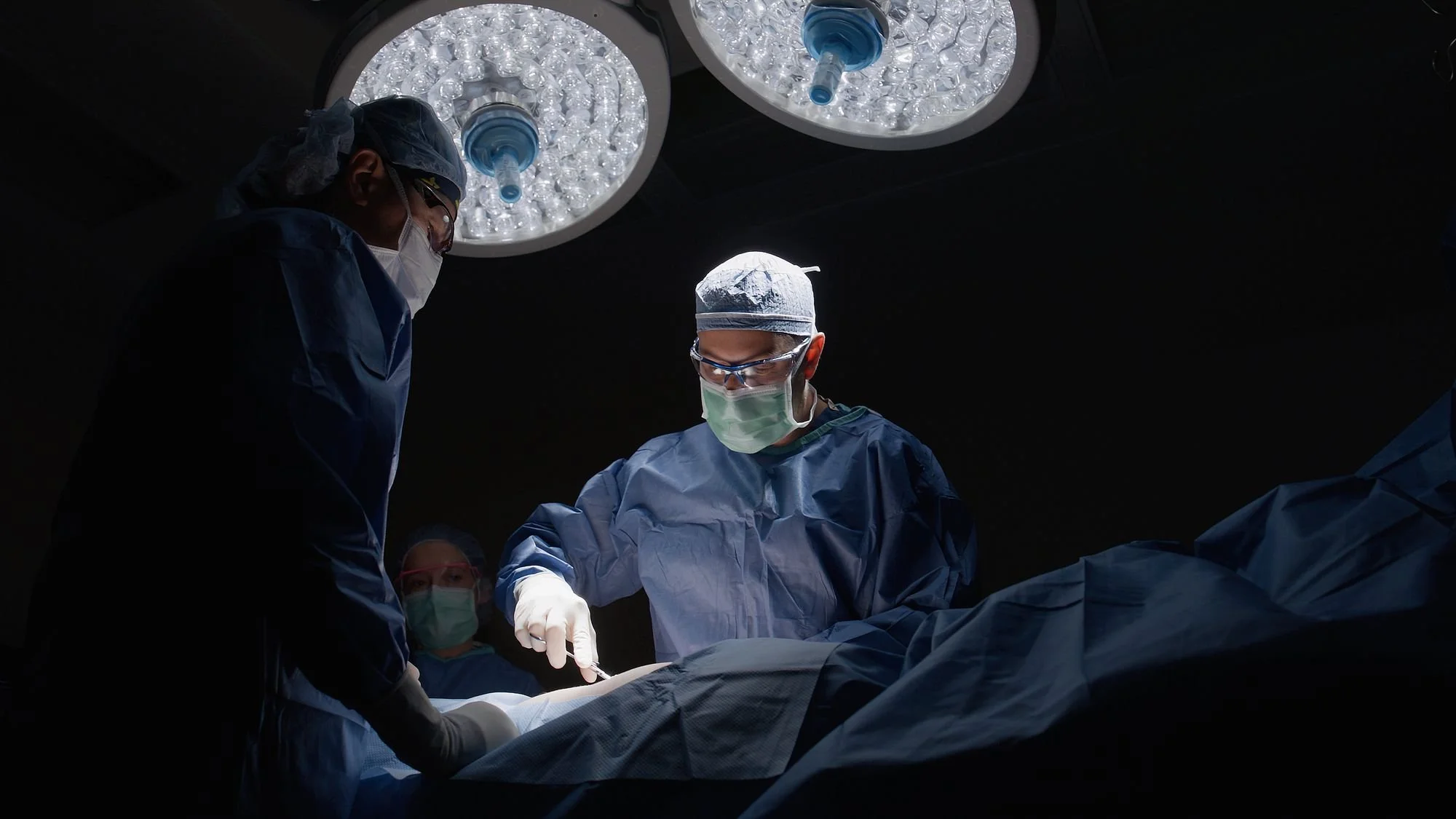 Medical professionals in surgical masks and gowns performing a surgery under bright overhead lights in an operating room.