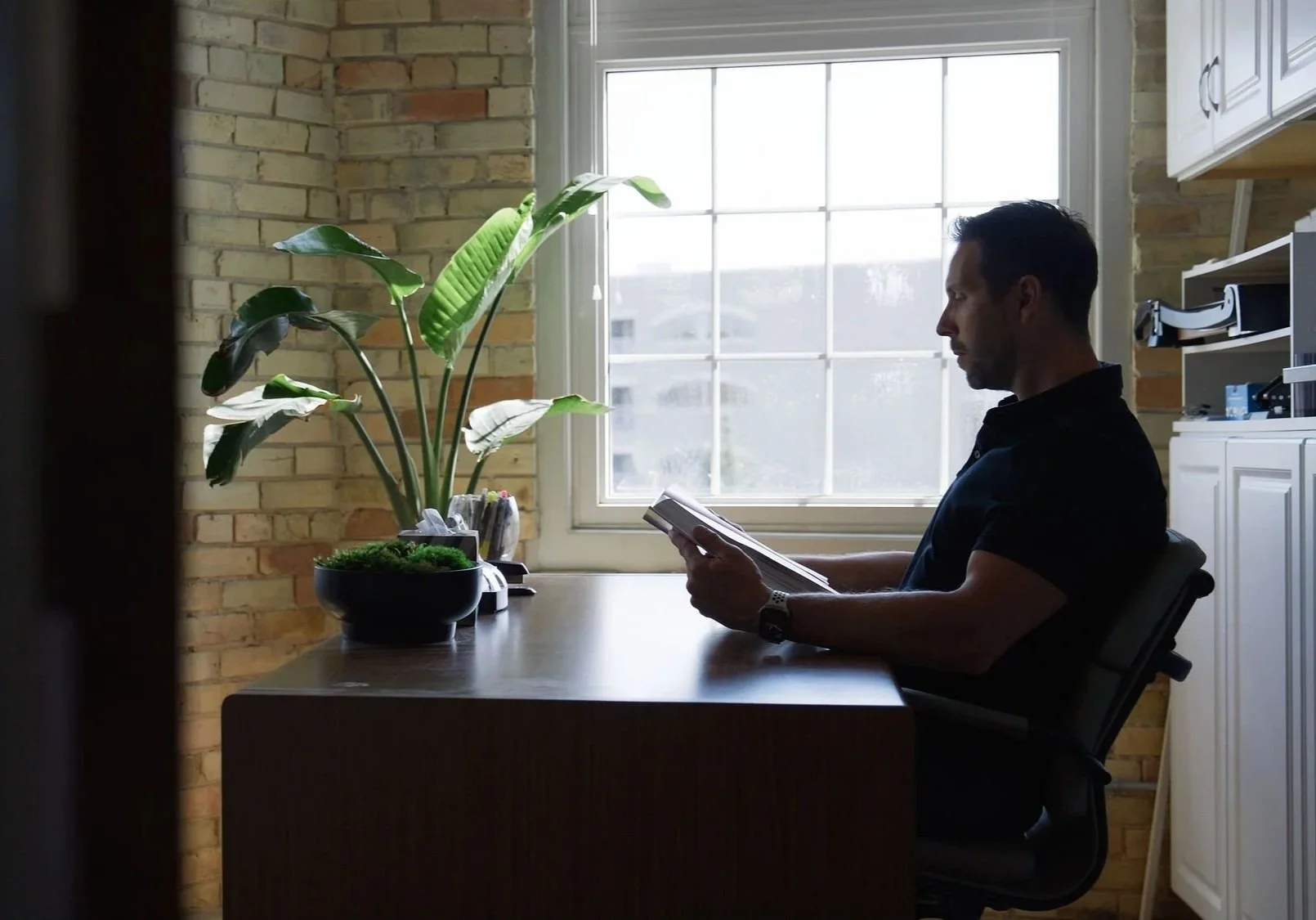 Man sitting at a desk, reading a book or magazine. Sunlight coming through large window behind him. Potted plant and office supplies on the desk. Brick wall in the background.