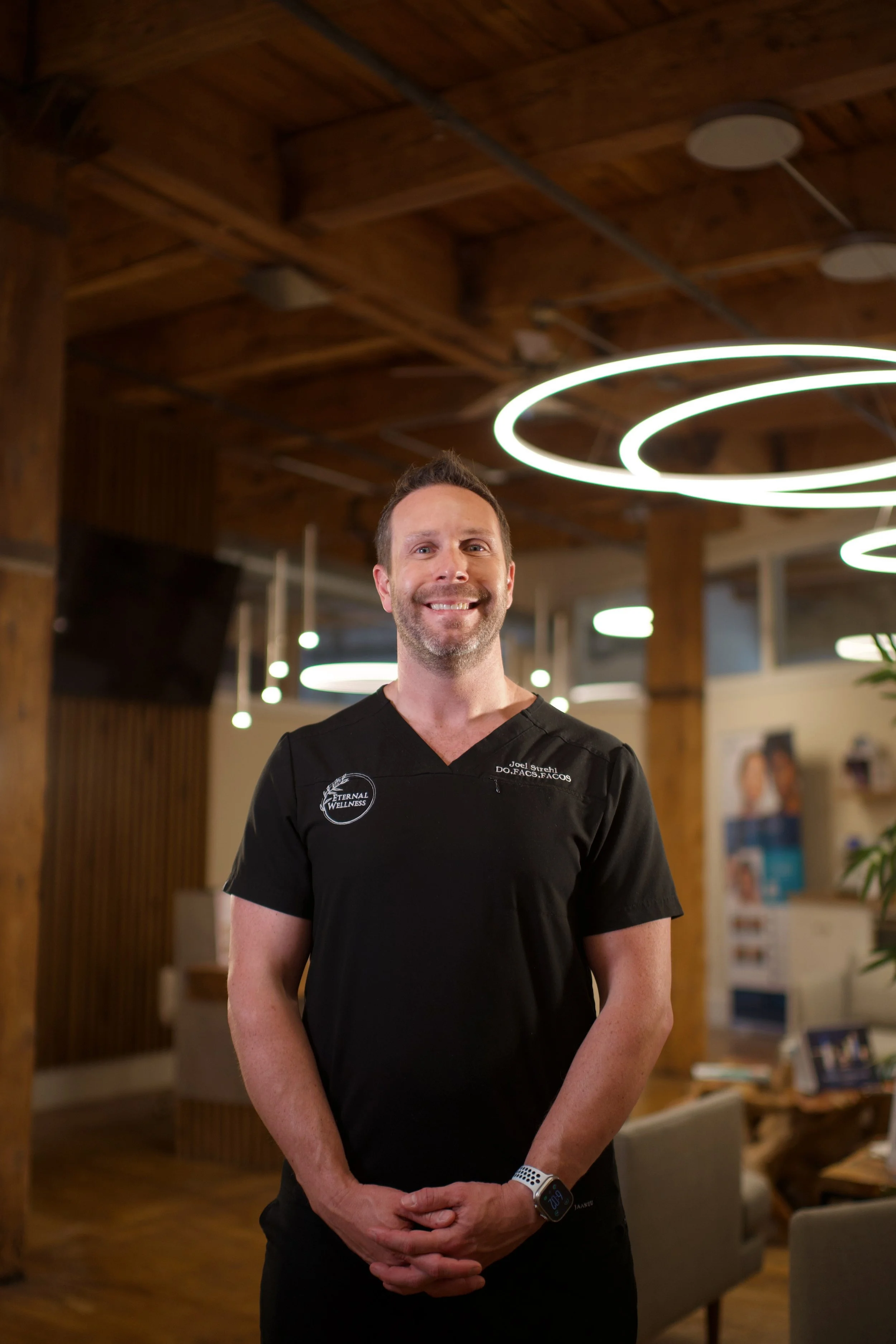 A smiling man in black medical scrubs standing indoors with a wooden ceiling and modern circular lighting fixtures in the background.