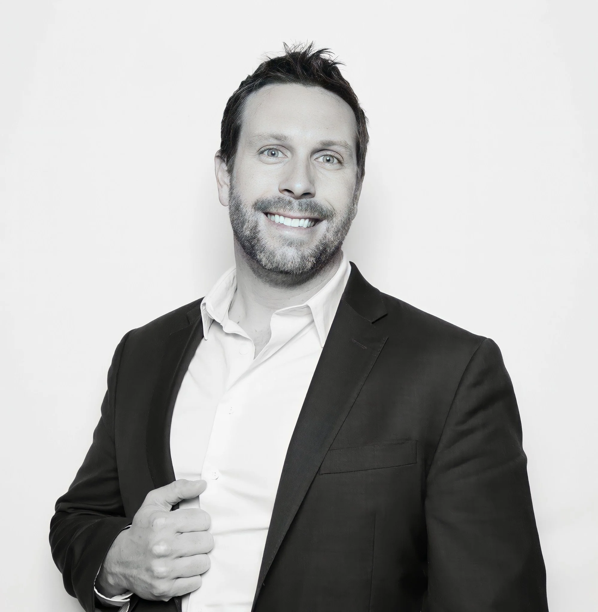 A man with a beard and mustache wearing a black tuxedo and white dress shirt, smiling, with his left hand pointing to his chest against a plain white background.