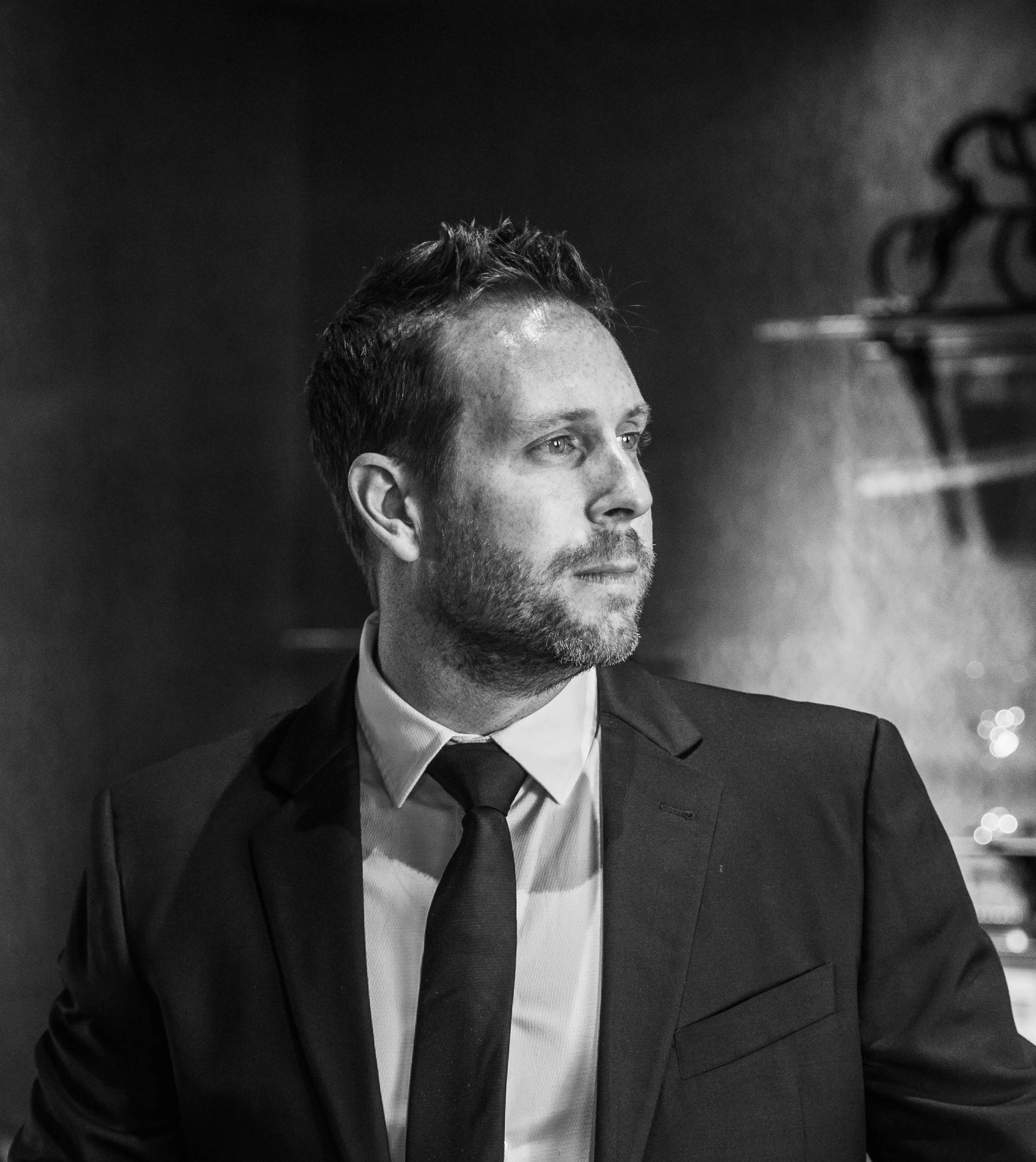 Black and white photo of a man wearing a suit, tie, and white shirt, with short hair and a beard, looking away.