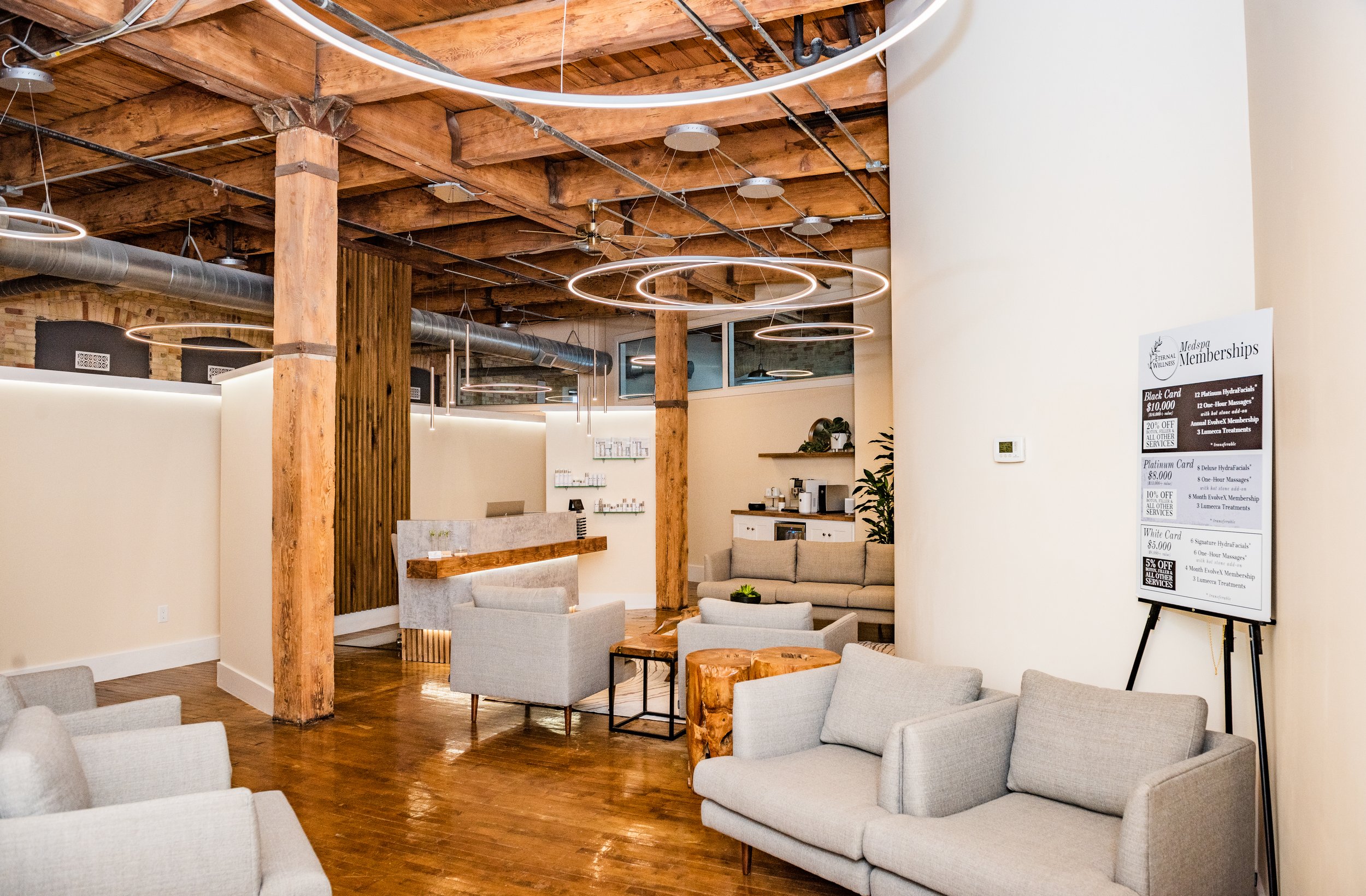 Modern waiting area with beige armchairs, wooden flooring, wooden support beams, and contemporary circular pendant lights.