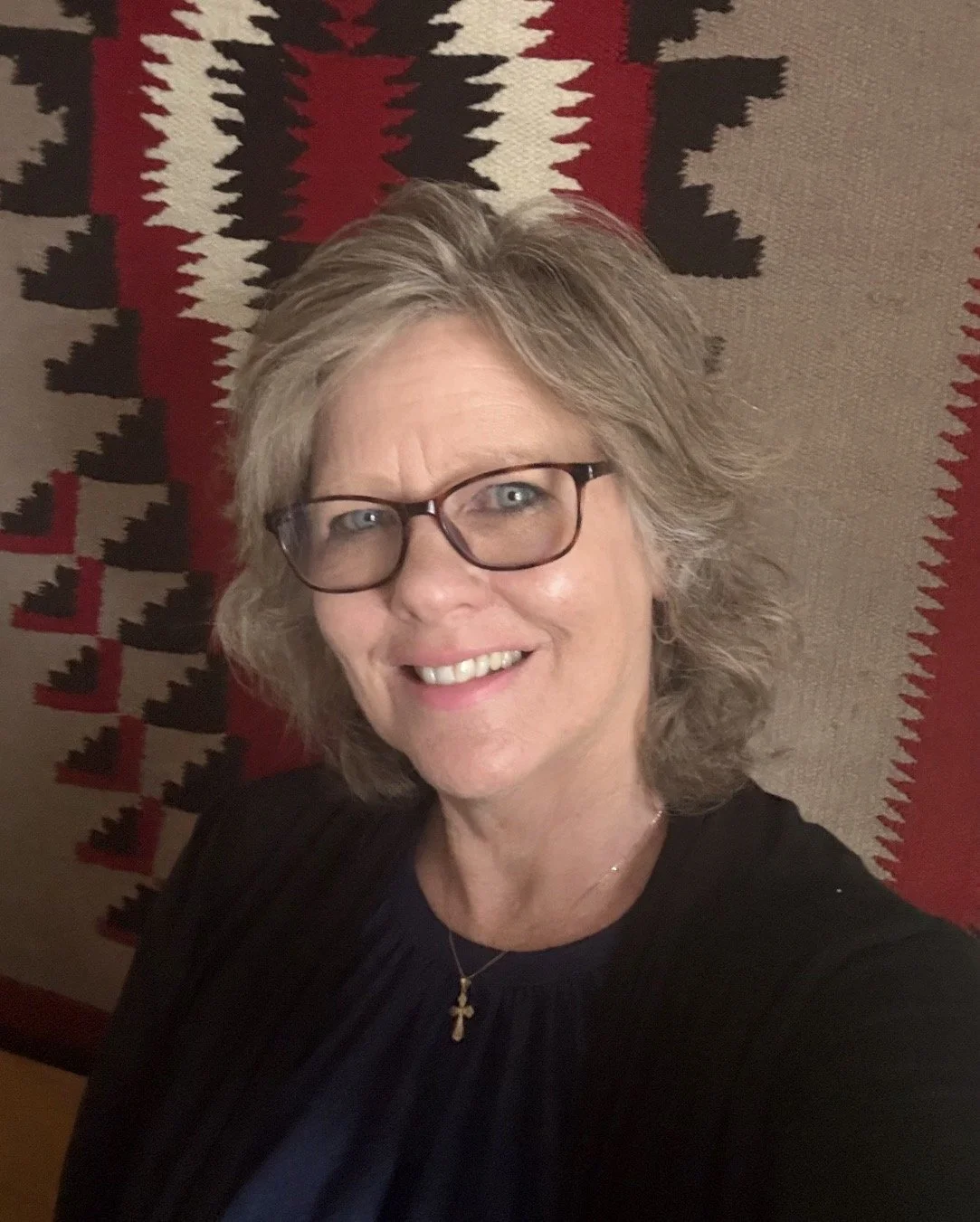 A woman with gray hair, glasses, and a smile, standing in front of a patterned red, black, and beige textile background.