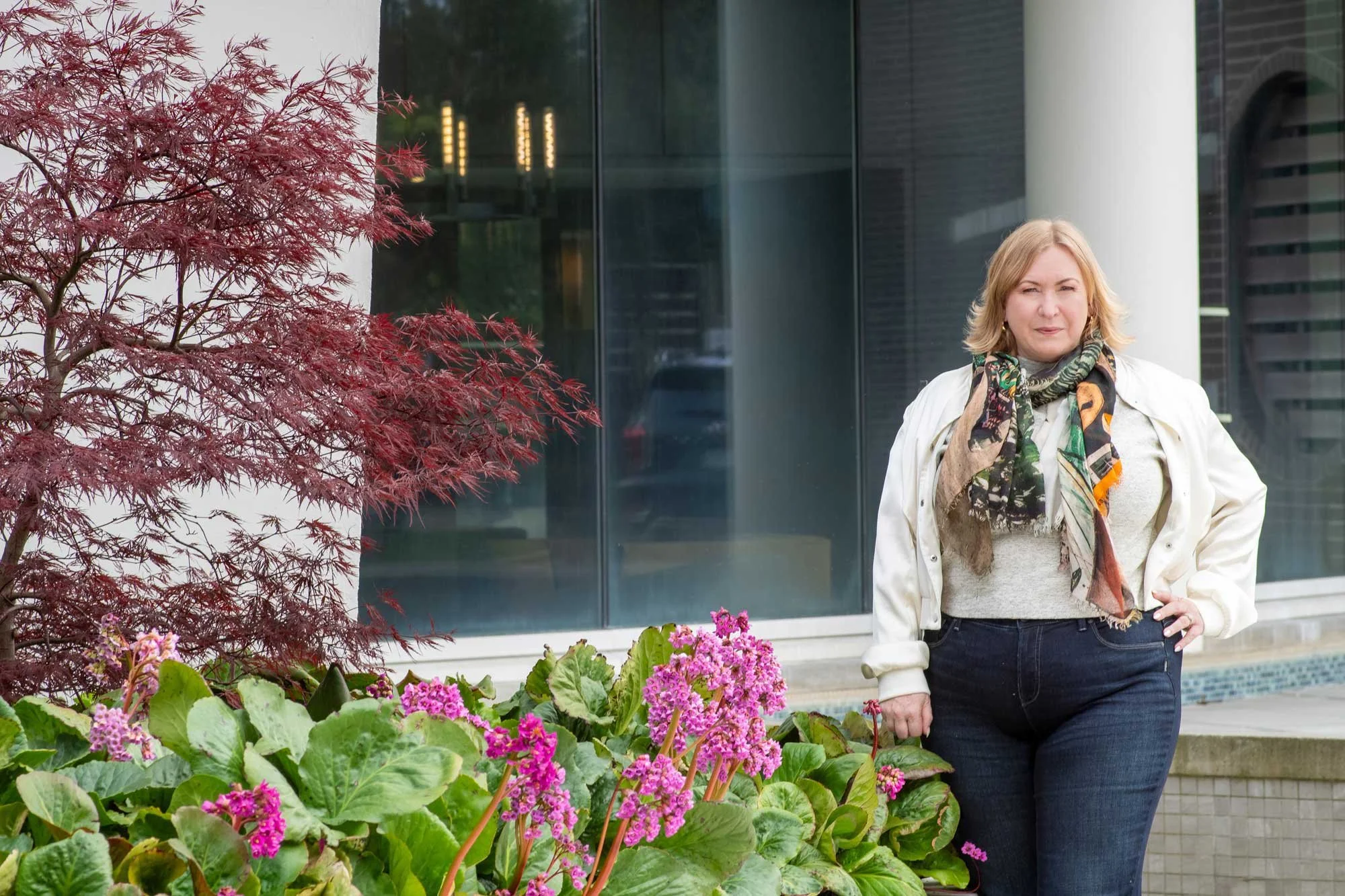 A woman with blonde hair wearing a white jacket, patterned scarf, and dark jeans standing outdoors near a modern building with large glass windows, surrounded by colorful plants and flowers, including a red-leaved tree and pink and purple flowers.