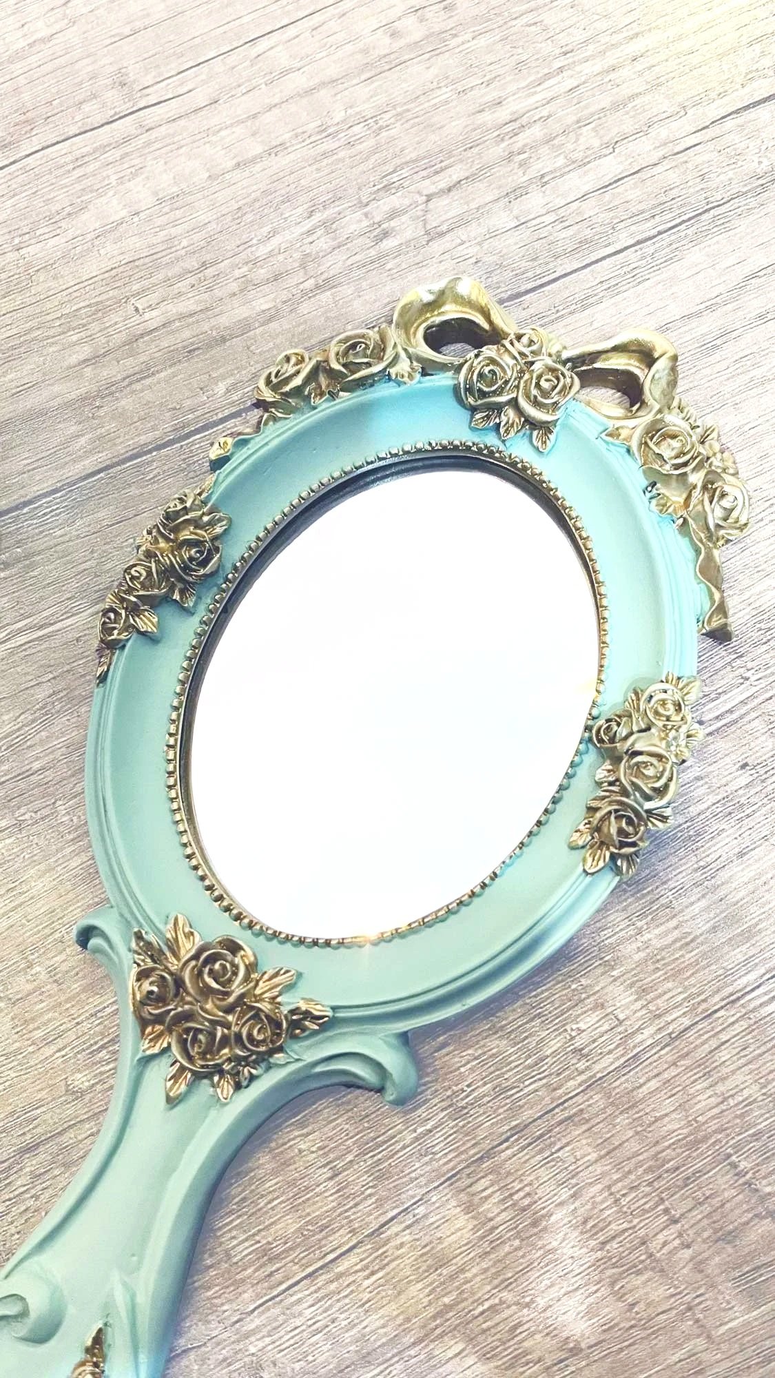 A vintage-style hand mirror with a light blue frame decorated with gold roses and leaves, lying on a wooden surface.