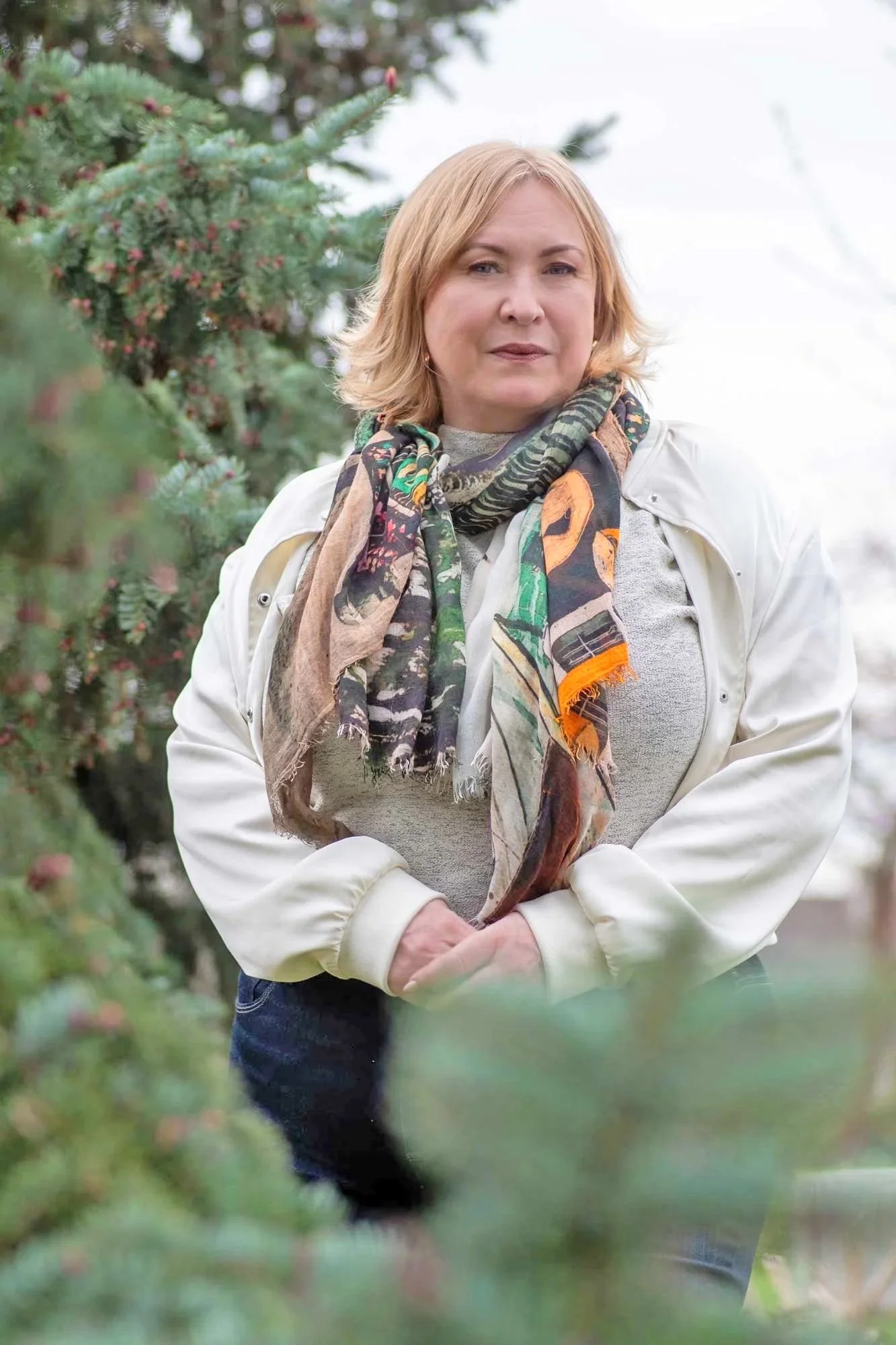 A woman with short, blonde hair, wearing a light-colored jacket and a colorful scarf, standing outdoors near green trees.