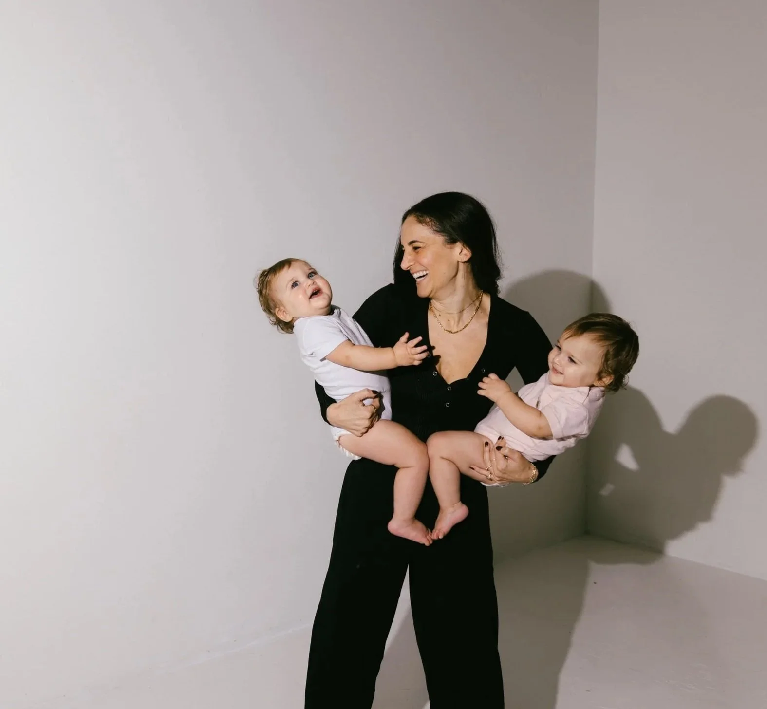 A woman in a black dress holding two young children, all smiling and laughing, against a plain white wall.