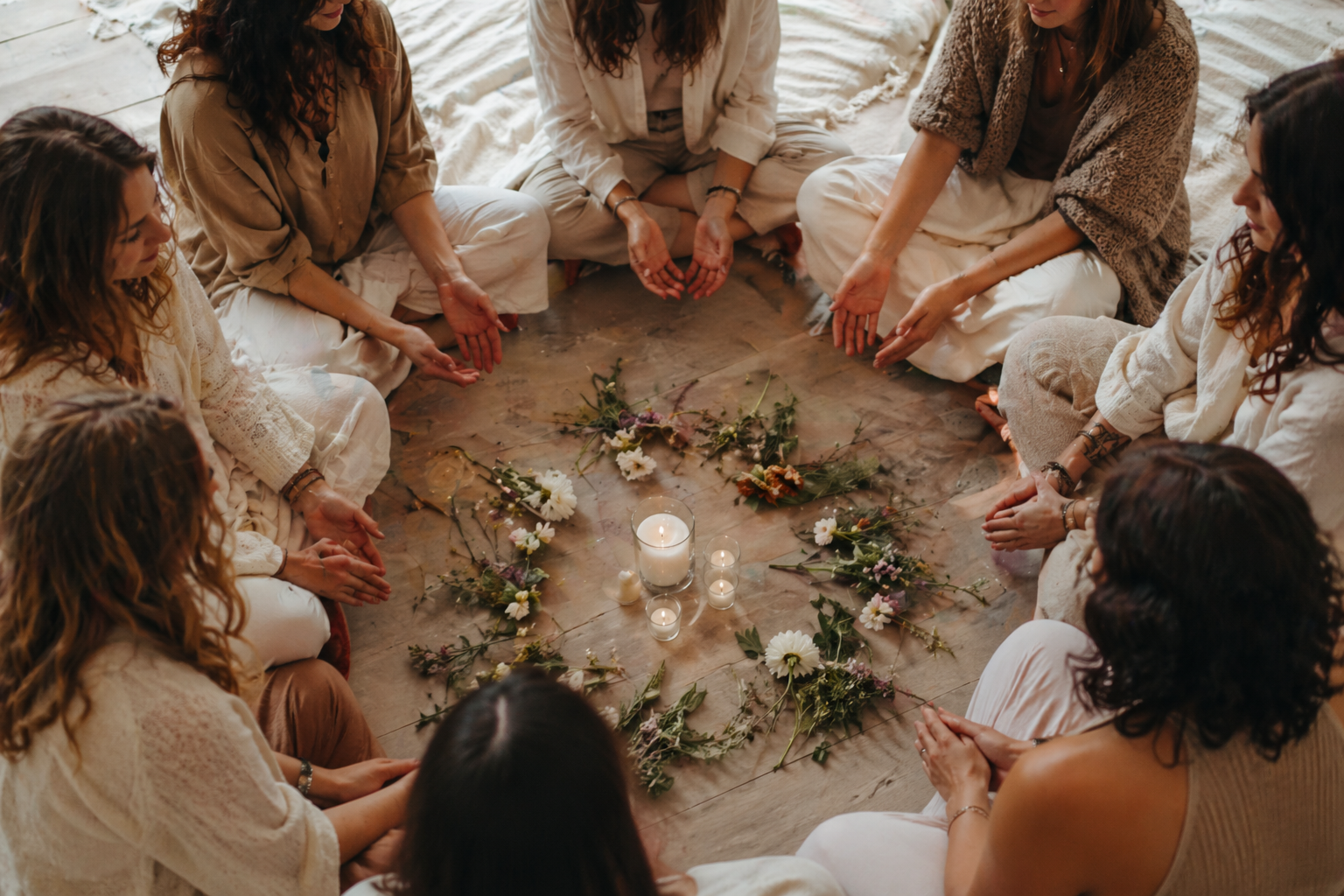 A group of women sitting in a circle on the floor with candles and flowers in the center, participating in a spiritual or meditation session.