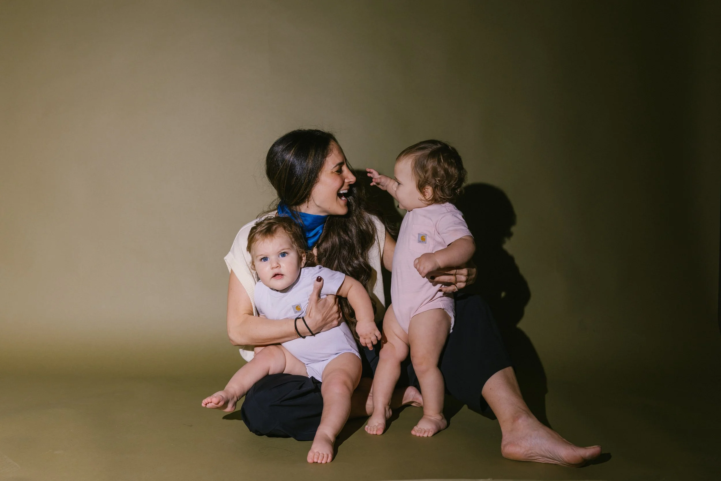 A woman with long dark hair holding two small children, one on each arm, against a plain background. The woman is smiling and interacting with a toddler girl, and the other child, a baby, is looking directly at the camera.