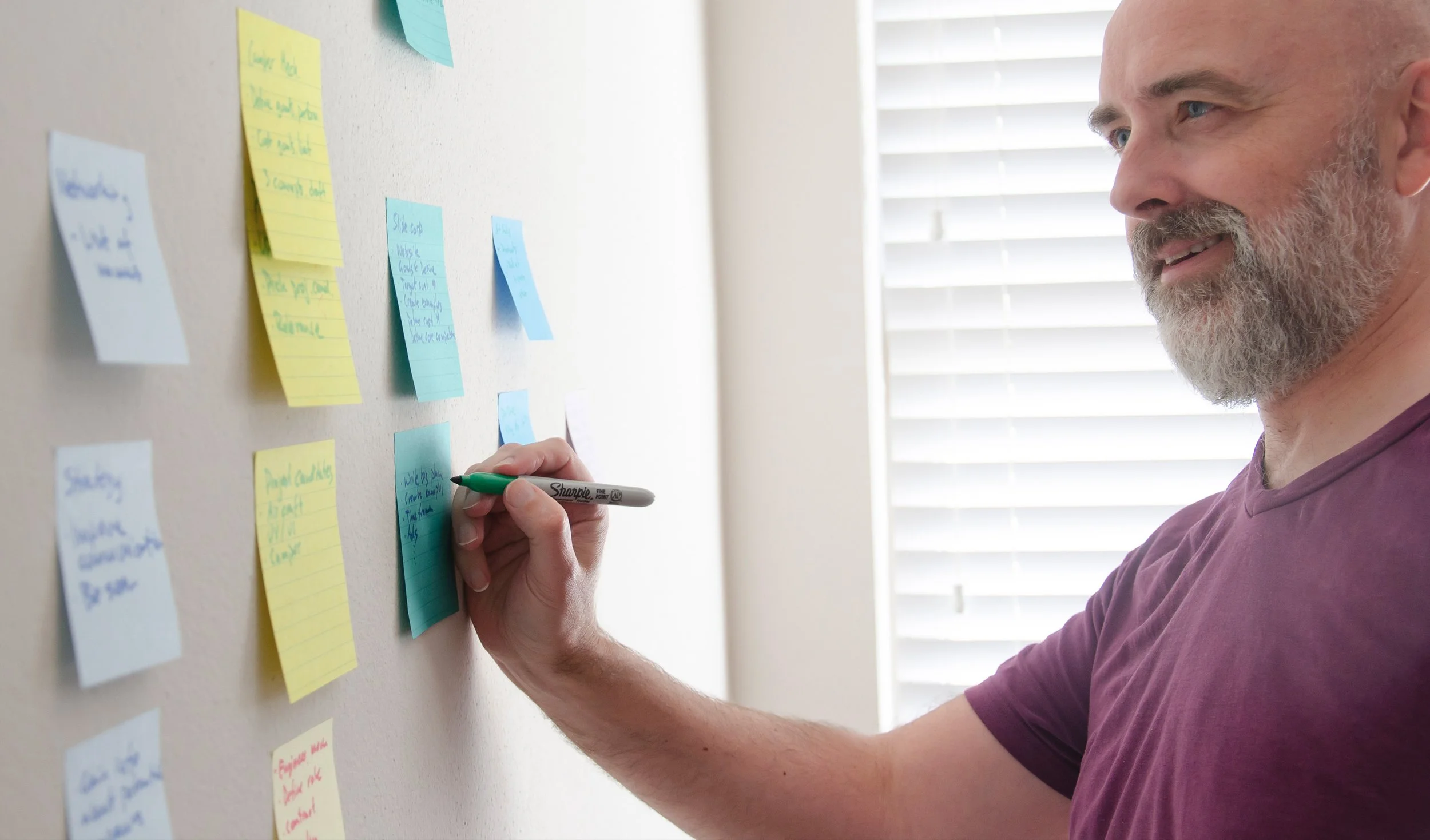 A man with a beard and mustache writing on sticky notes on a wall, using a green marker. The sticky notes are in various colors and contain handwritten notes, with a window with blinds in the background.