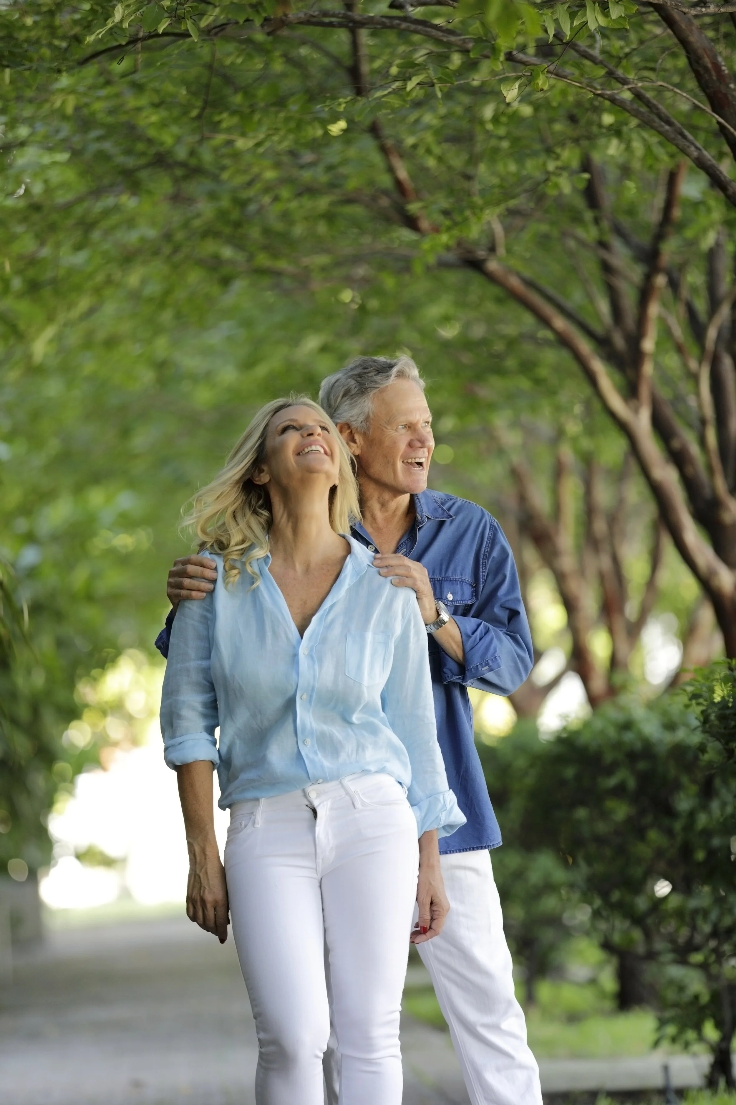 An older romantic couple enjoying a walk outdoors, smiling and looking up, surrounded by green trees.