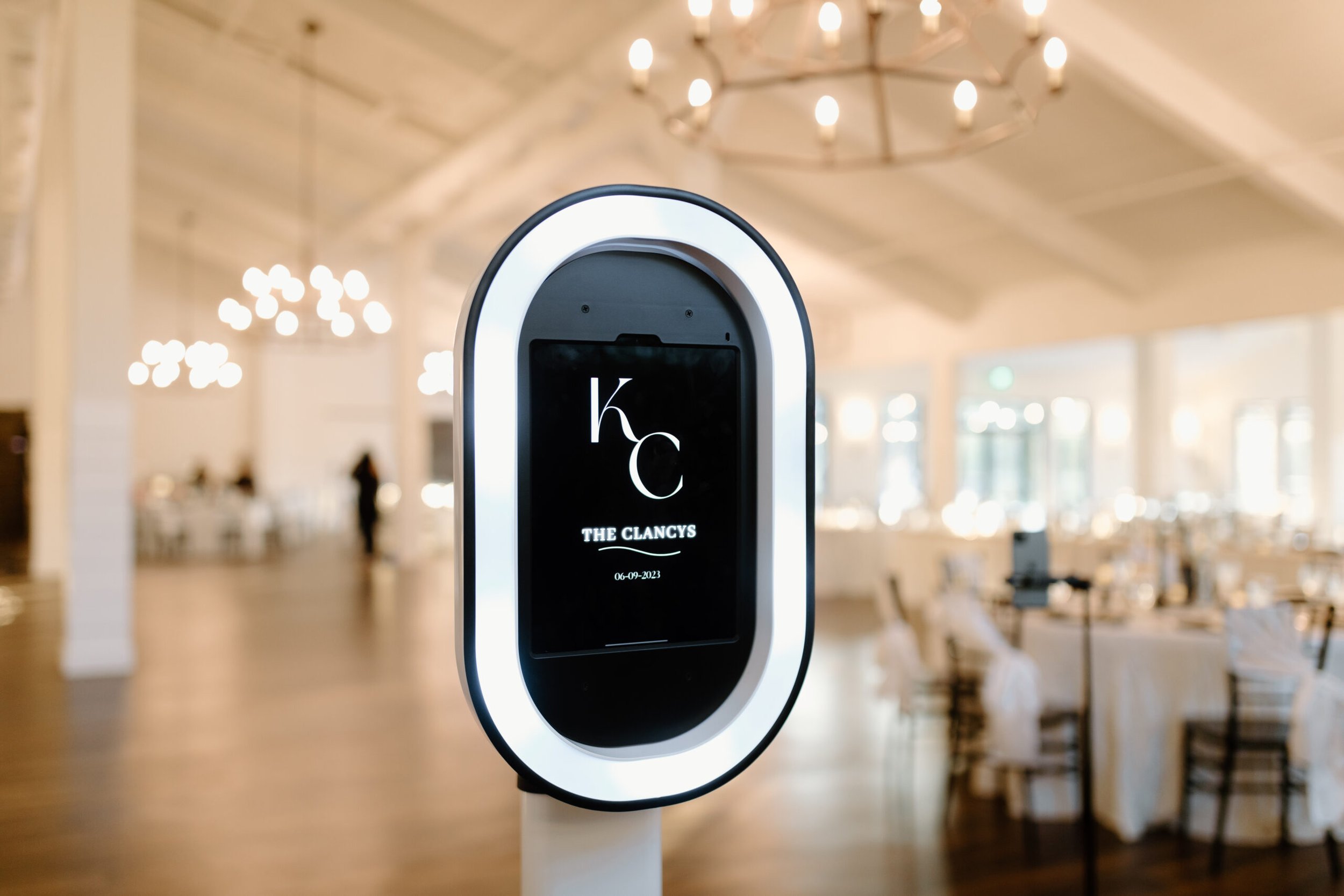 Digital sign with black background and white text displaying the words 'The Clancys' and the date '06-09-2023', mounted on a white stand in a spacious, well-lit interior with hanging lights and blurred seating area in the background.