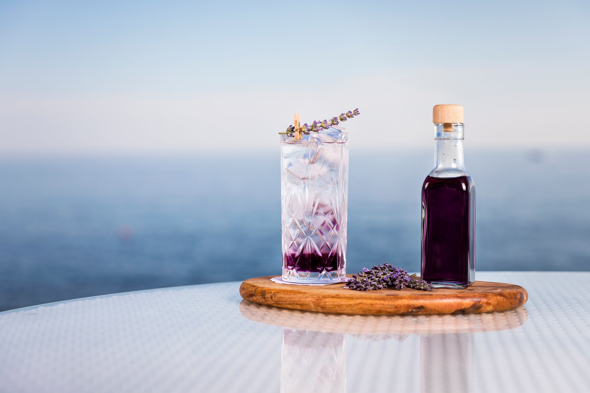A tall glass with a pink drink and lavender sprig garnish, and a small glass bottle with a purple liquid, both on a wooden board with lavender flowers in front, against a blurred blue background.