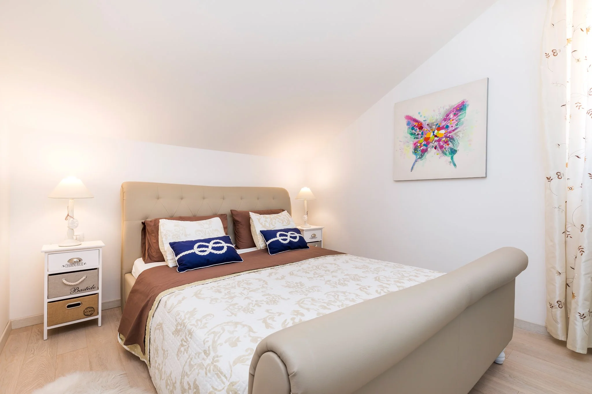 A cozy bedroom with a white upholstered bed, decorative pillows, a white nightstand with storage drawers, two lamps on either side, a colorful butterfly painting on the wall, and a window with floral curtains.
