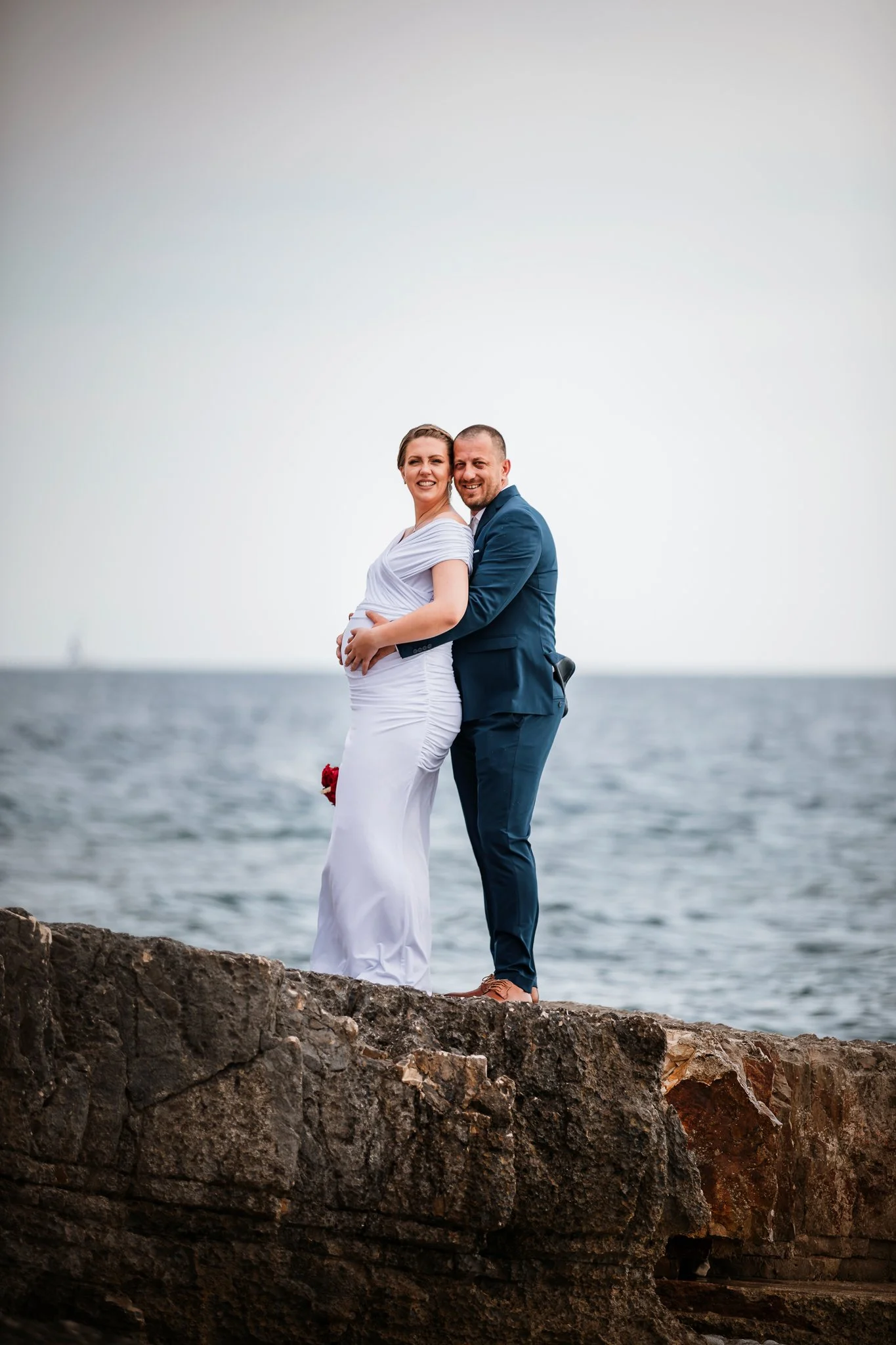 A smiling couple, the woman visibly pregnant, standing on a rocky shoreline by the water. The woman wears a white dress and the man is in a blue suit. They are embracing and smiling at the camera.
