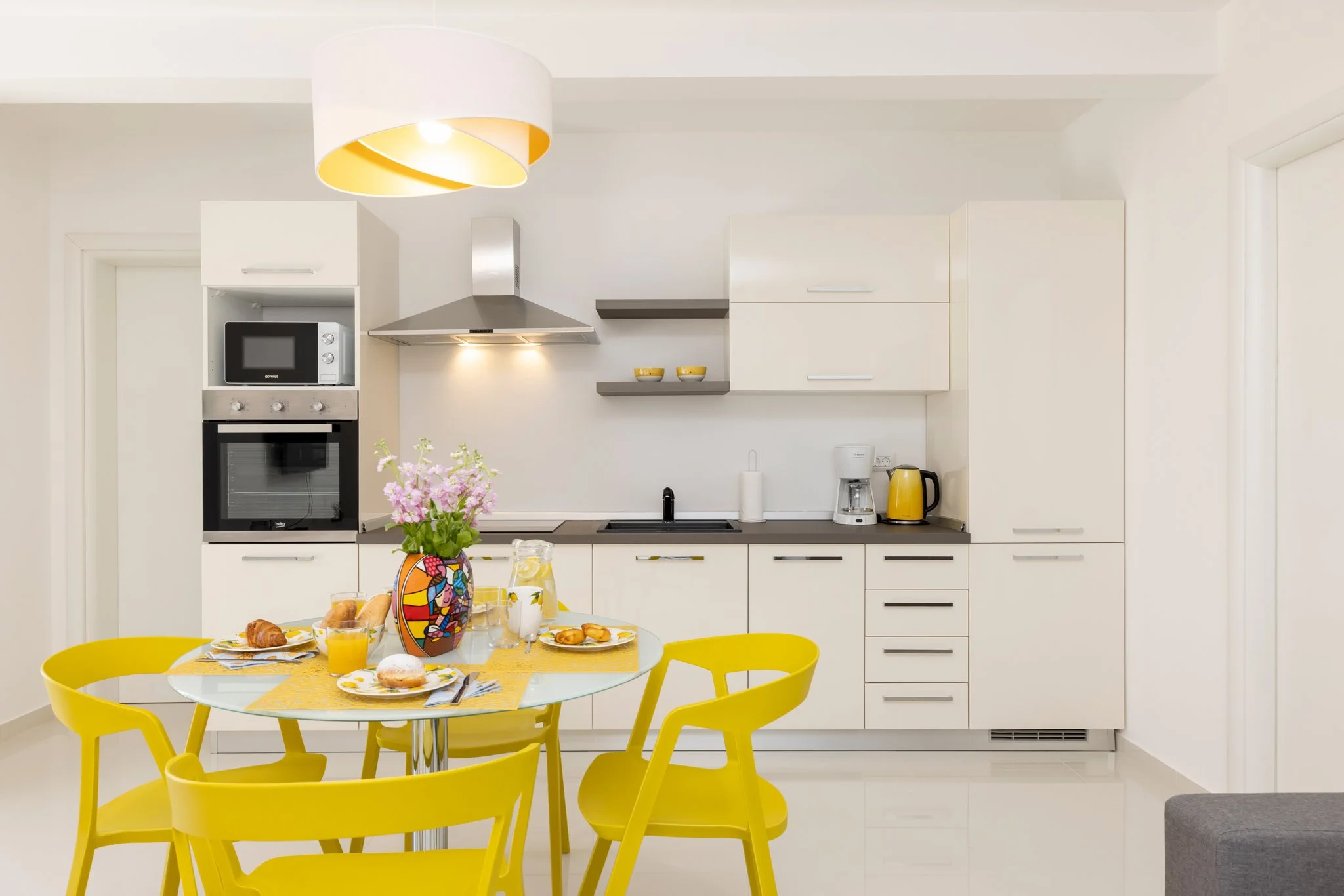 A modern kitchen with white cabinets, stainless steel appliances, and a black sink. A yellow dining table with six matching yellow chairs is set with plates, glasses, croissants, and orange juice. A vase with pink and purple flowers decorates the tab