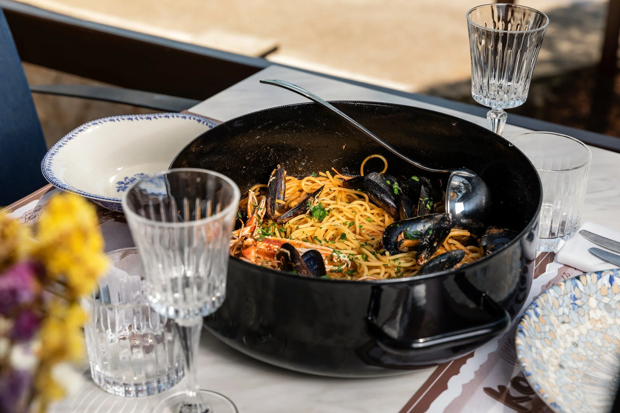 A table set with a large black bowl of spaghetti with mussels and herbs, empty glassware, and a decorative plate, with a blurred background and some flowers in the foreground.