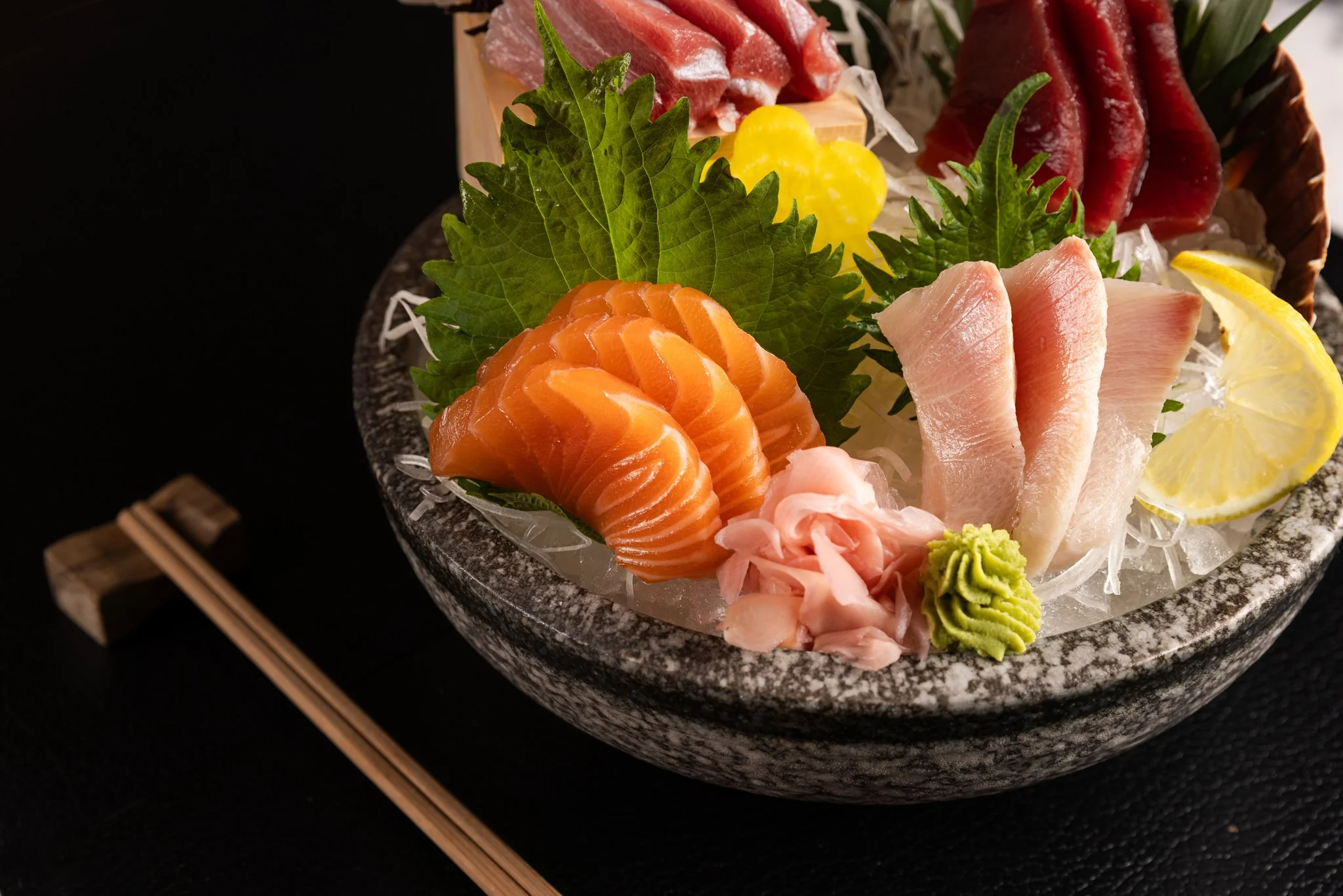 Assorted sashimi, including salmon, tuna, and white fish, arranged on a black, stone serving platter with green shiso leaves, lemon slices, wasabi, and shredded daikon garnish.