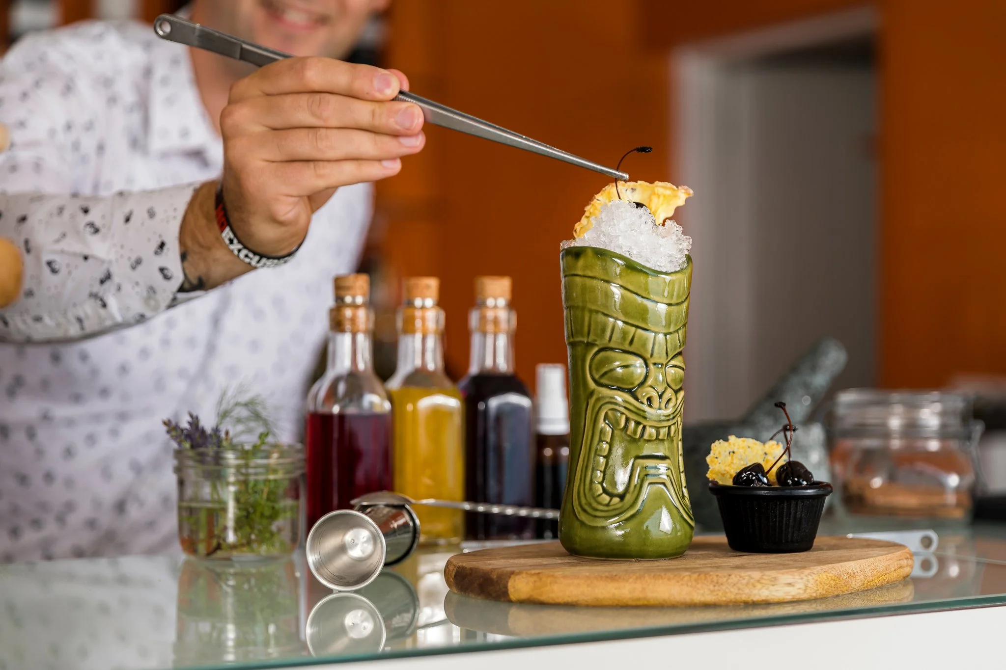 Person smiling as they prepare a tropical cocktail in a tiki mug, surrounded by various ingredients and bottles on a kitchen counter.