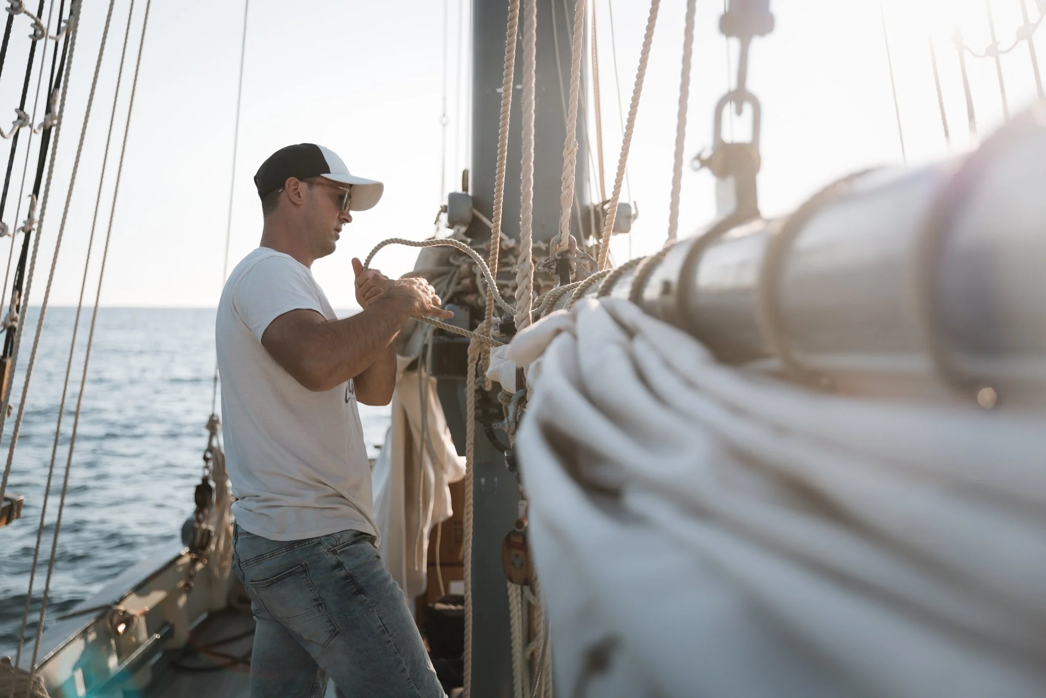 A man wearing a white t-shirt, jeans, sunglasses, and a baseball cap working on the rigging of a sailboat at sea during sunset.