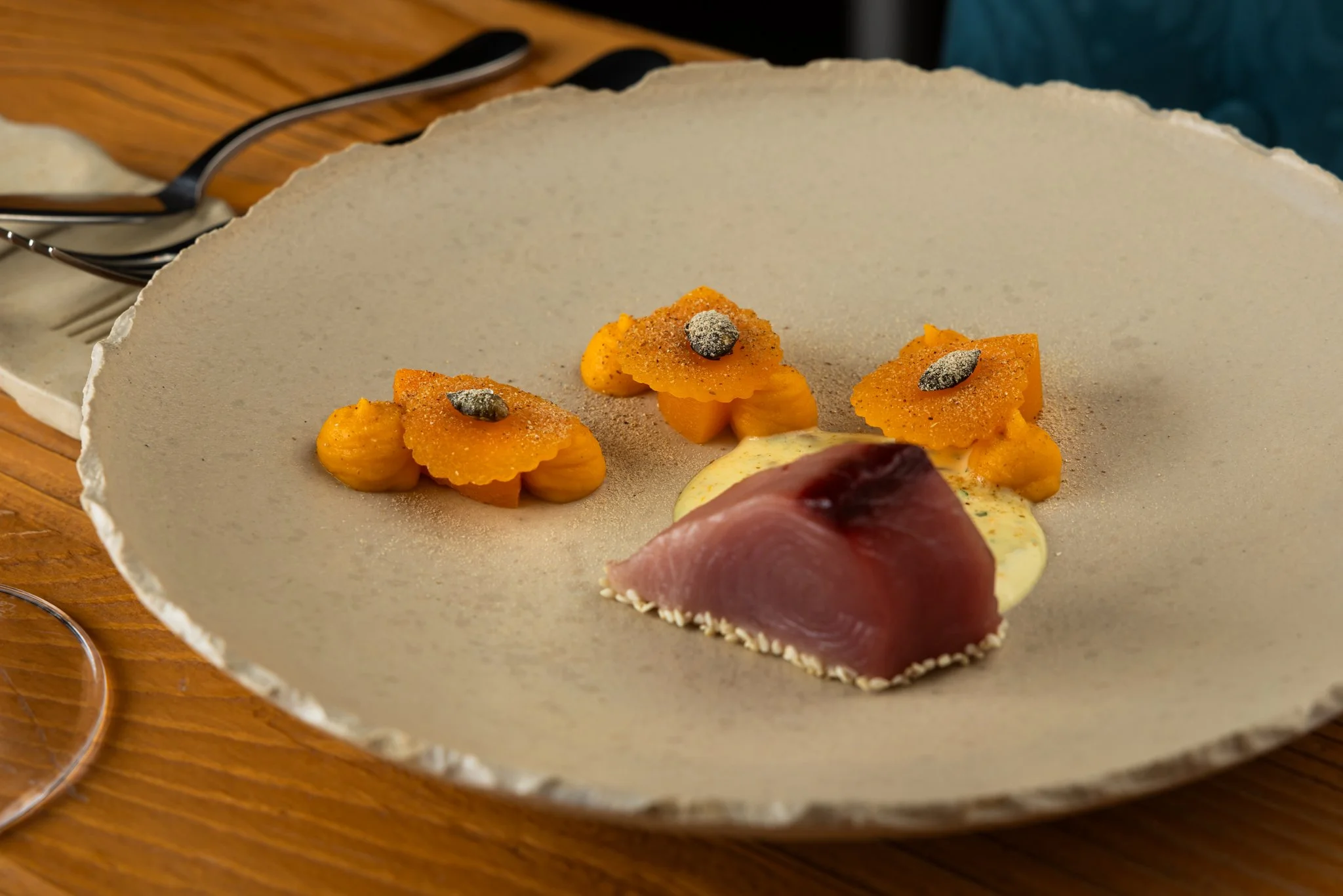 A close-up of a plate with three pieces of sliced butternut squash garnished with black pepper and a small dollop of yellow sauce, with a slice of cured meat, possibly prosciutto, on the side, on a round textured ceramic plate.