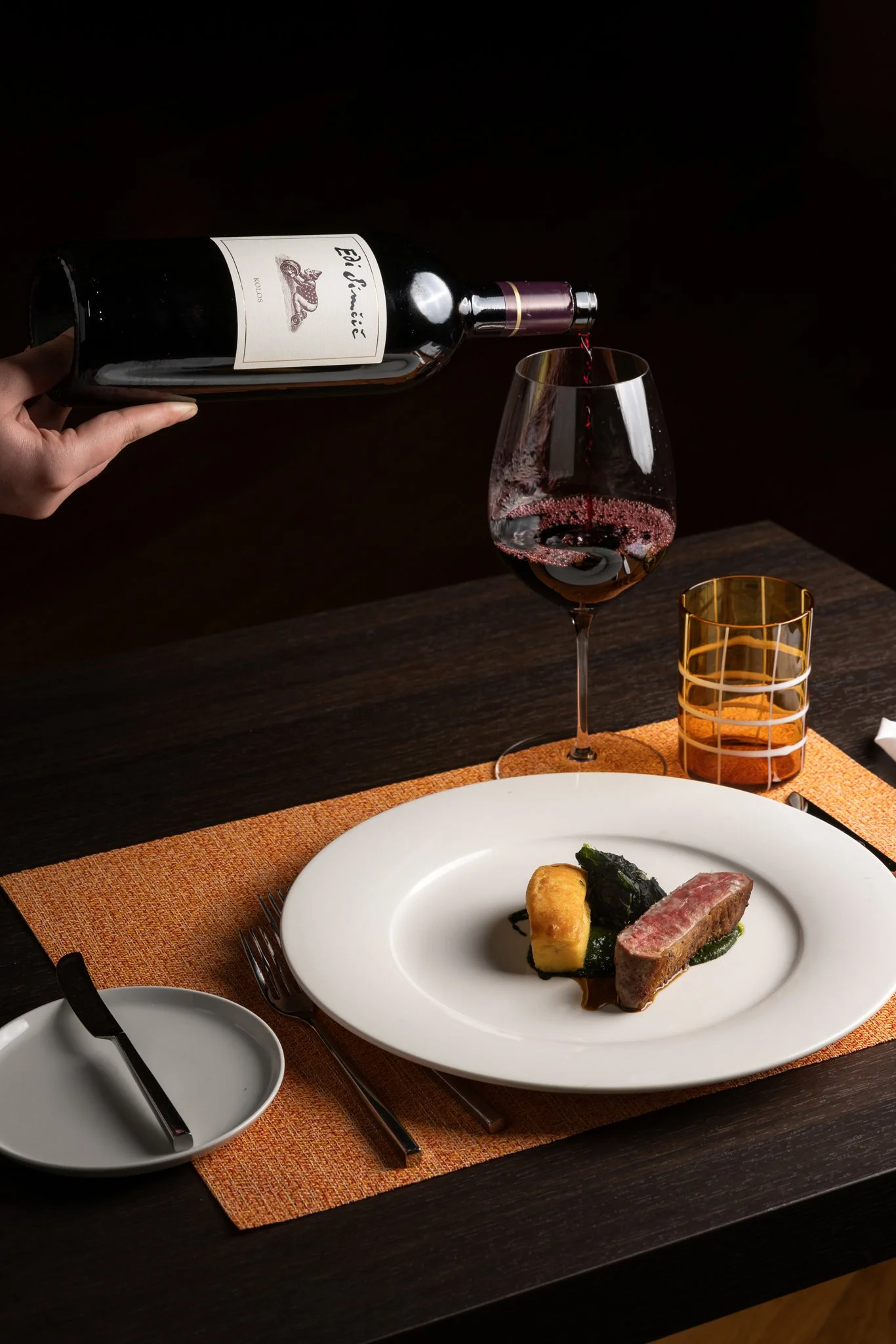 A hand pouring red wine into a wine glass on a dining table set with a plate of food, a small white dish, a fork, and a knife. The table has an orange placemat.