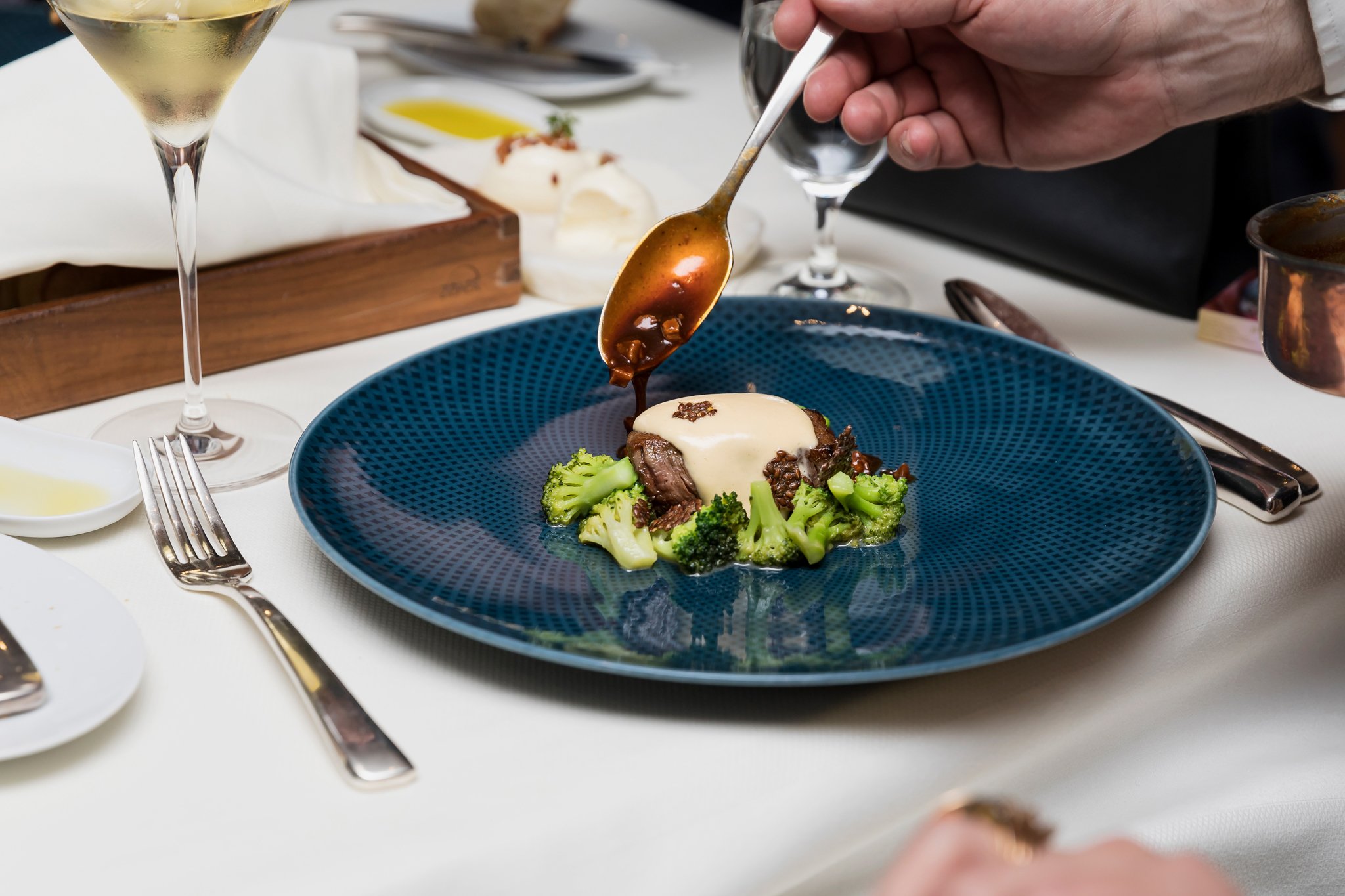 A plated dish of beef topped with sauce, served with broccoli, on a blue plate, with a hand pouring sauce from a spoon. A glass of white wine and other tableware are also visible.