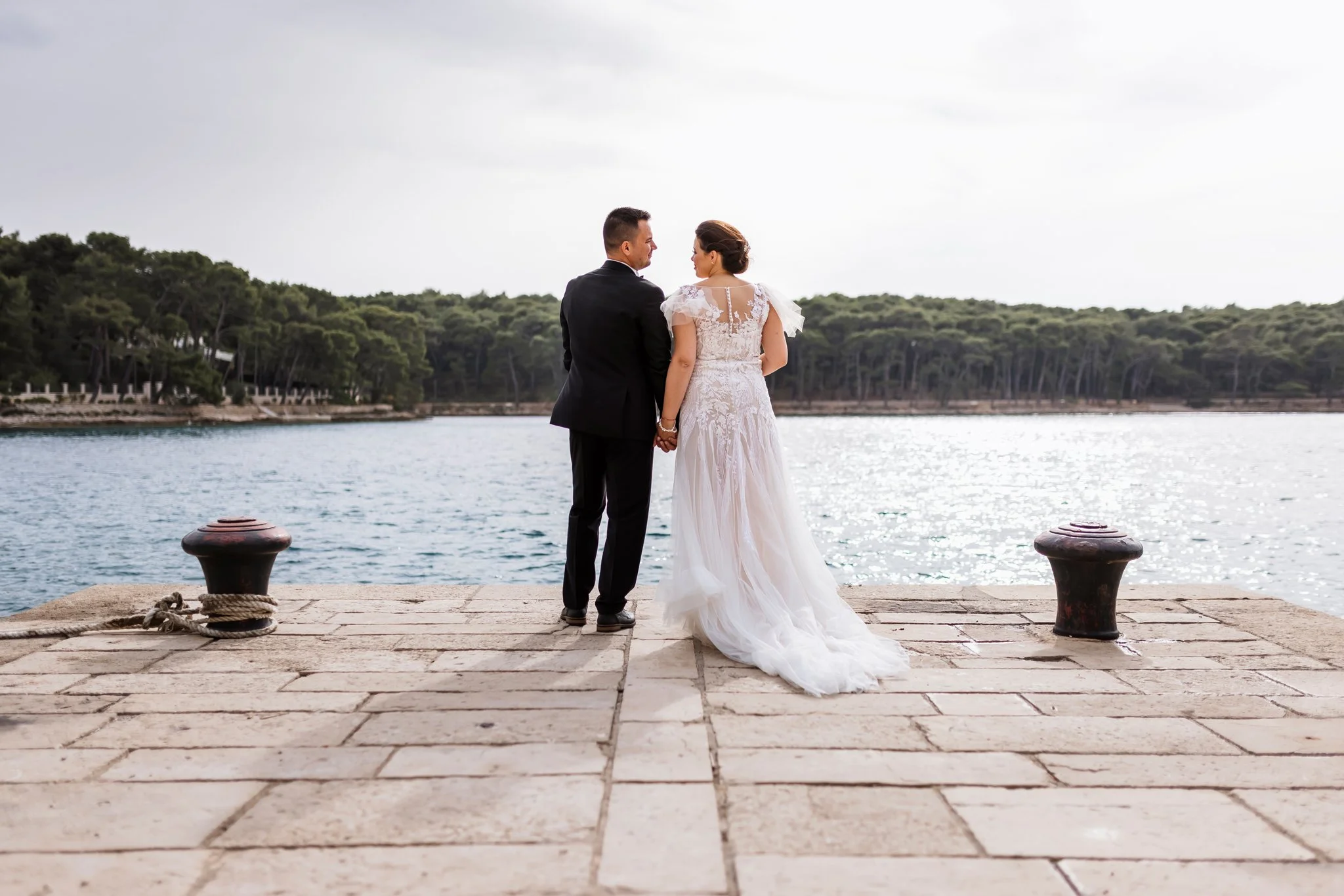 A bride and groom holding hands and looking at each other near a body of water with trees in the background. They are standing on a stone-paved dock with mooring bollards.