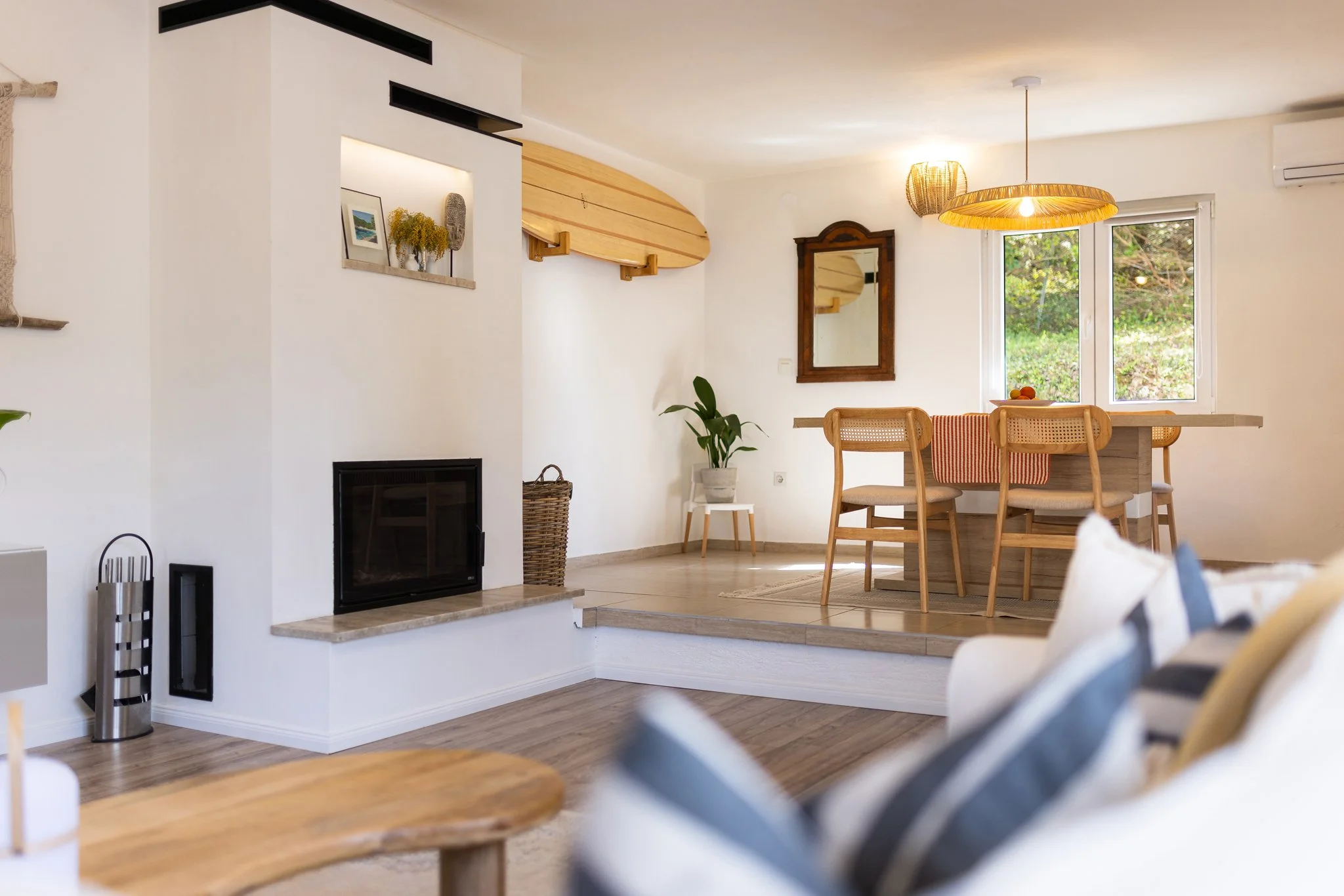 Interior view of a modern living room and dining area with white walls, a fireplace, wooden furniture, decorative items, and a window showing greenery outside.