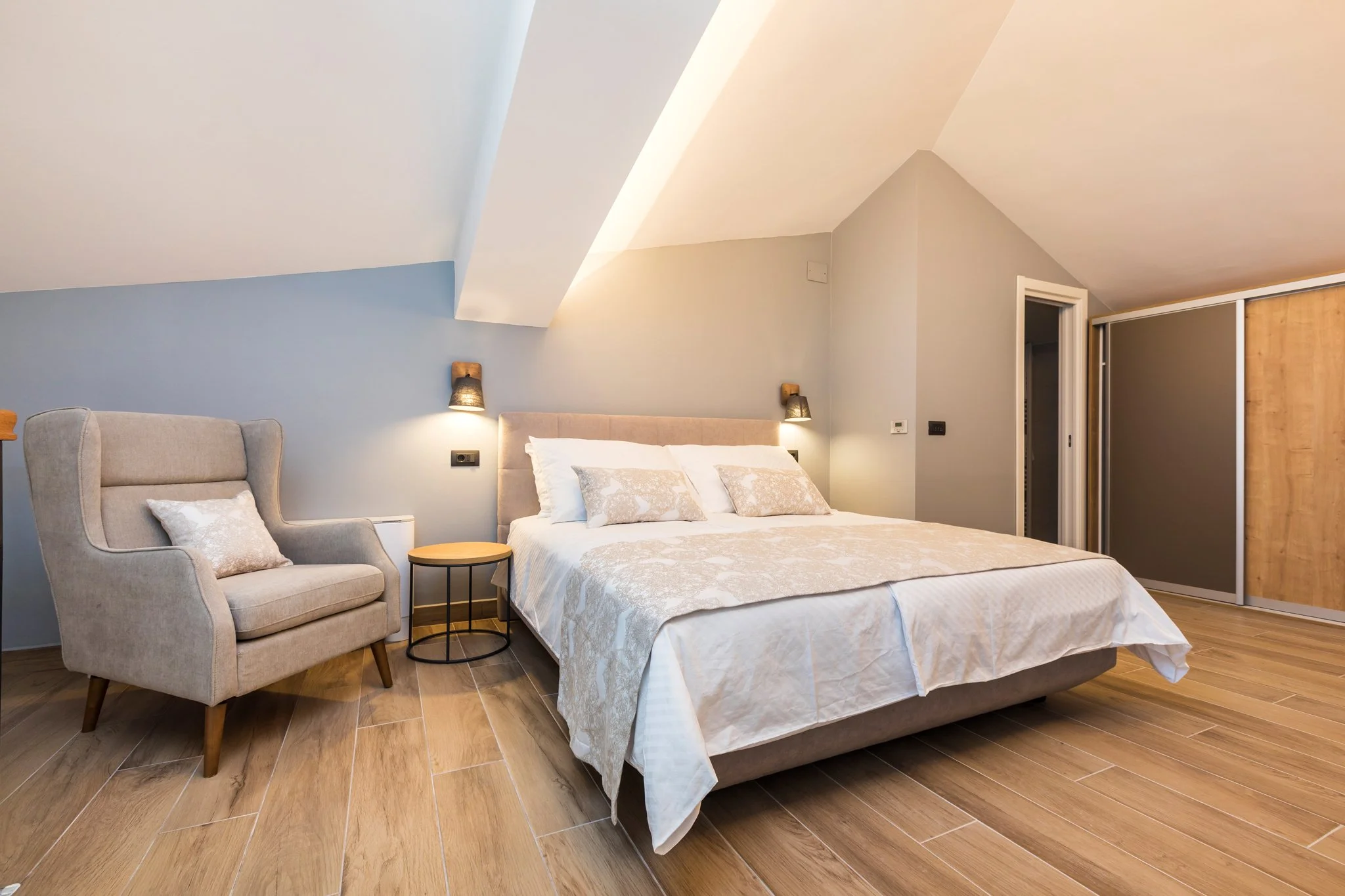 A bedroom with a beige upholstered bed, white bedding and throw pillows, a beige armchair with wooden legs and a cushion, and wooden flooring. The walls are painted in light colors with two wall-mounted lamps above the bed, and there is a sliding doo